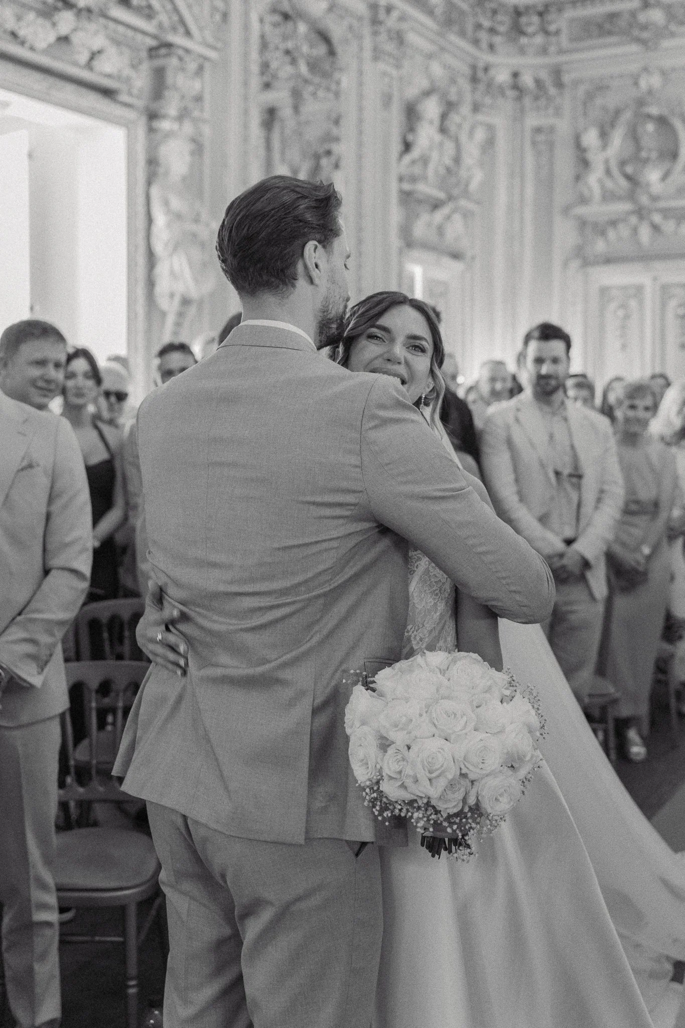 A bride and groom are dancing during their wedding reception. The bride is holding a bouquet of white roses and is smiling at the groom. Guests are watching and smiling in the background, in an ornate room with detailed walls and ceiling decor.