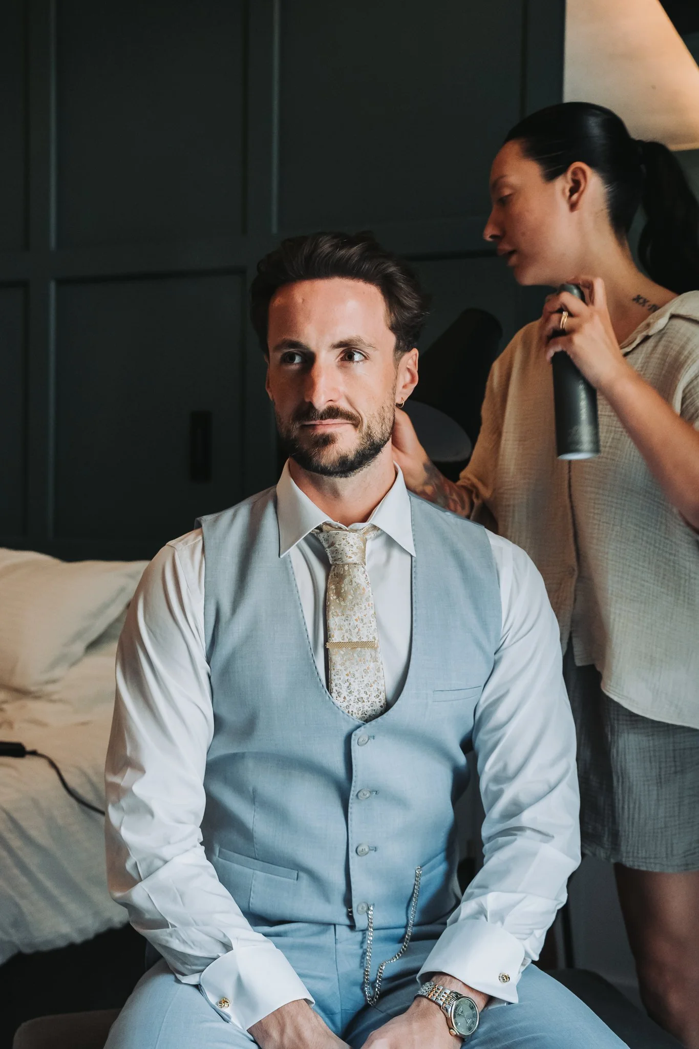 A man dressed in a light gray vest, shirt, and tie sitting on a bed, while a woman with dark hair in a ponytail styles his hair with a spray bottle in her hand.