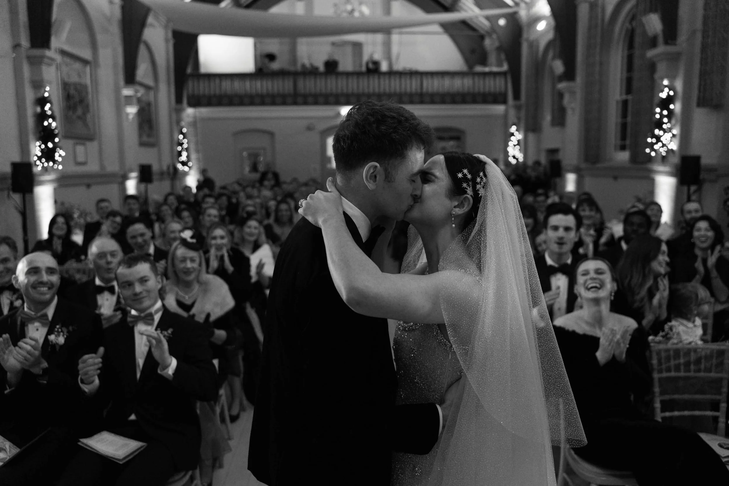 A black and white photo of a bride and groom kissing during their wedding ceremony, surrounded by seated guests in a decorated indoor venue.