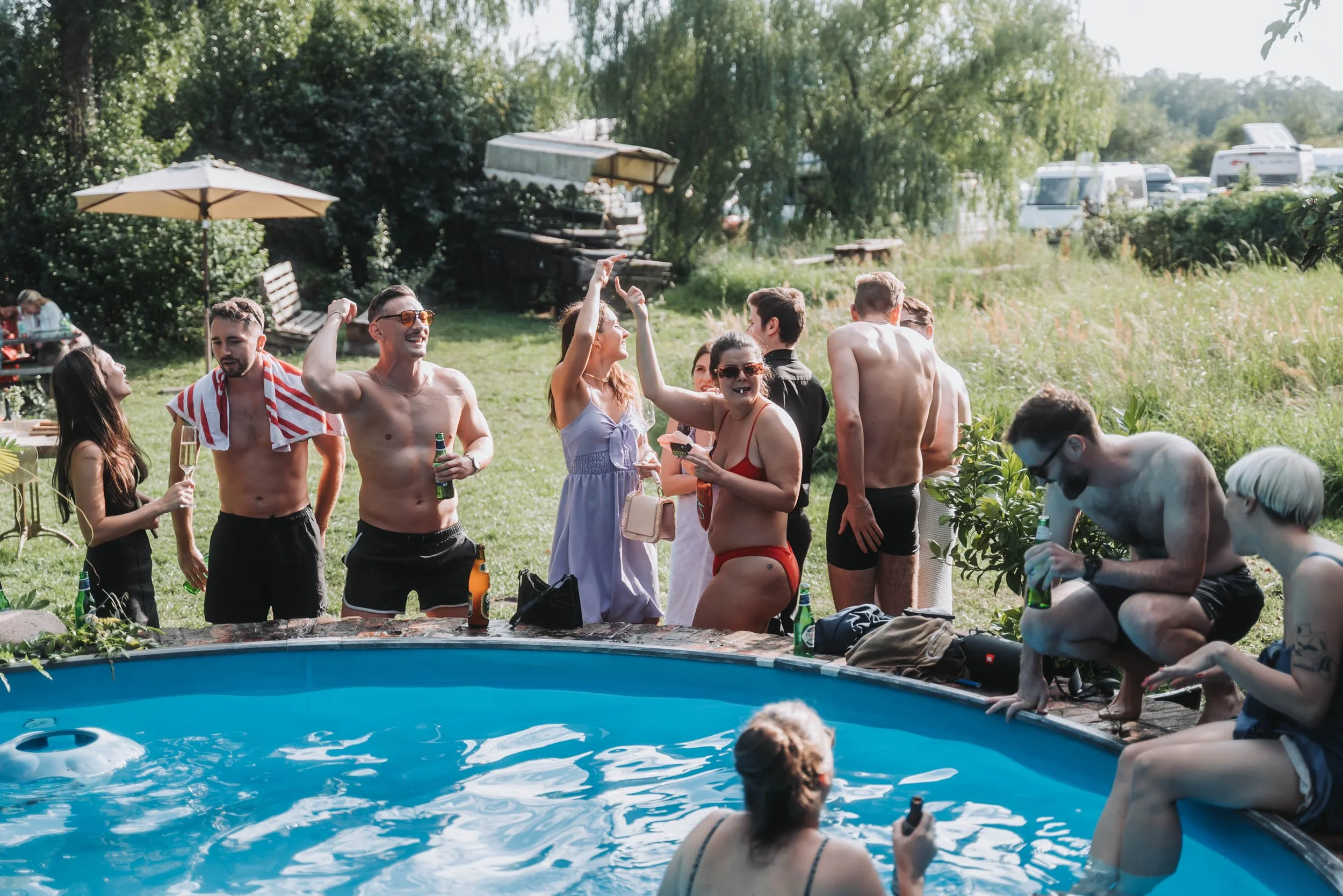 Group of people at an outdoor pool party, some in swimsuits, standing next to the pool, holding drinks, and socializing.