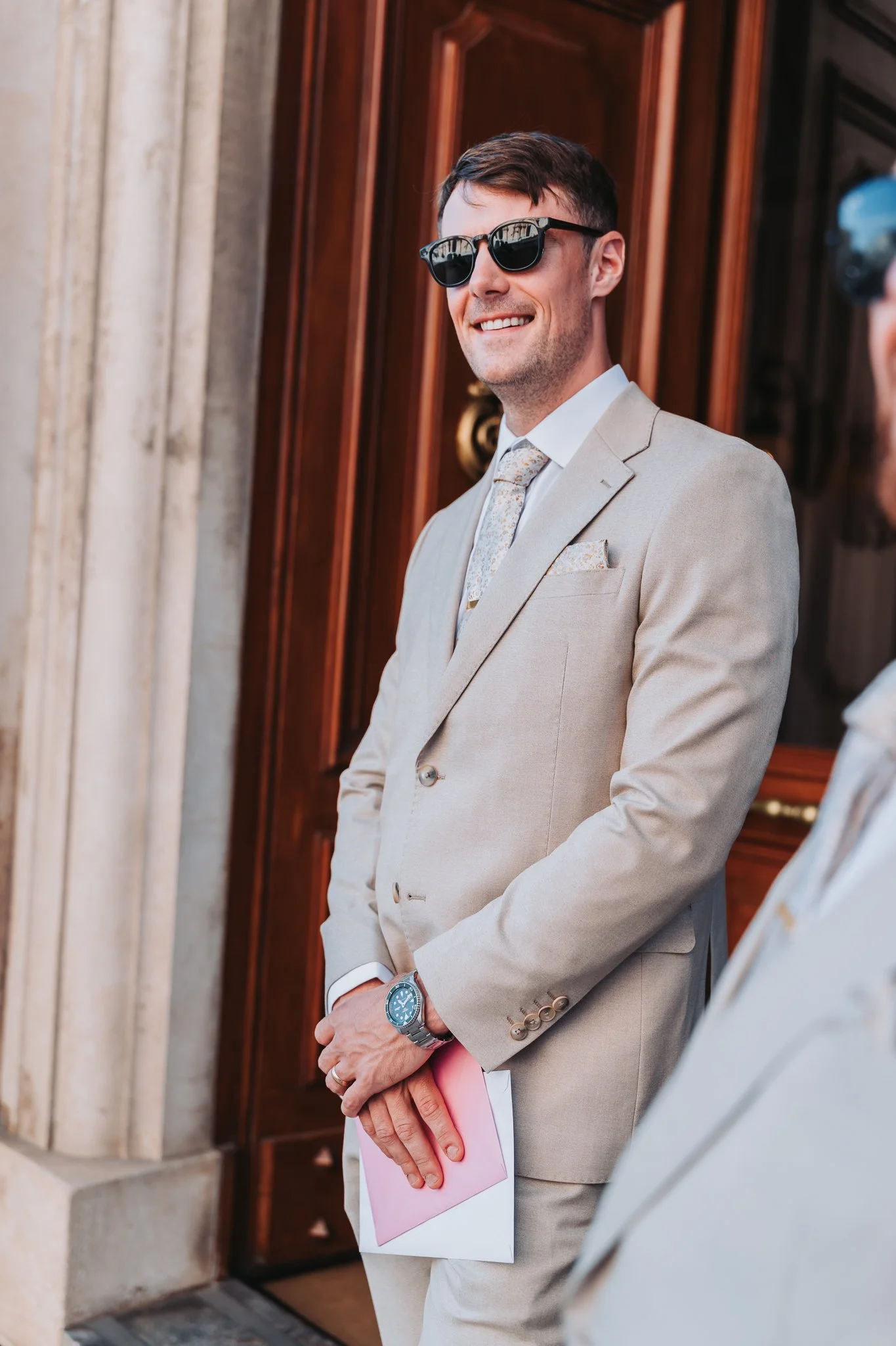 A man dressed in a beige suit with a patterned tie, wearing sunglasses, smiling and holding pink papers, standing outside a building with wooden doors and stone columns.