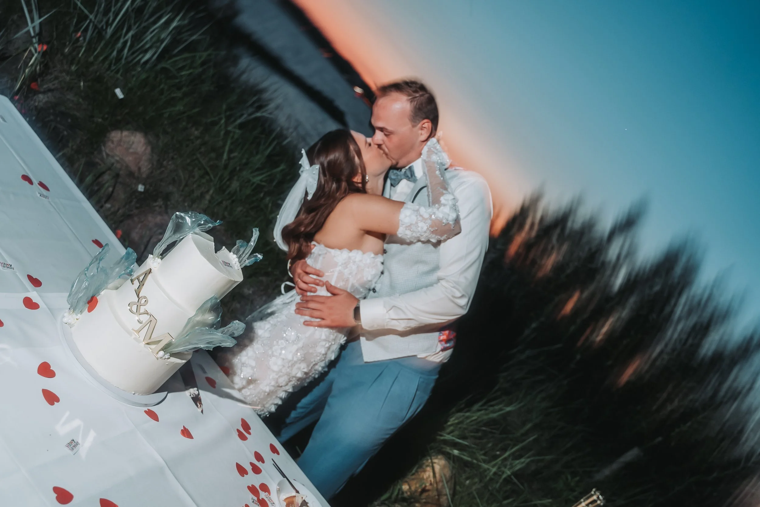 A bride and groom kissing outdoors during sunset, celebrating their wedding with a decorated cake on a table covered with a white cloth with red hearts.