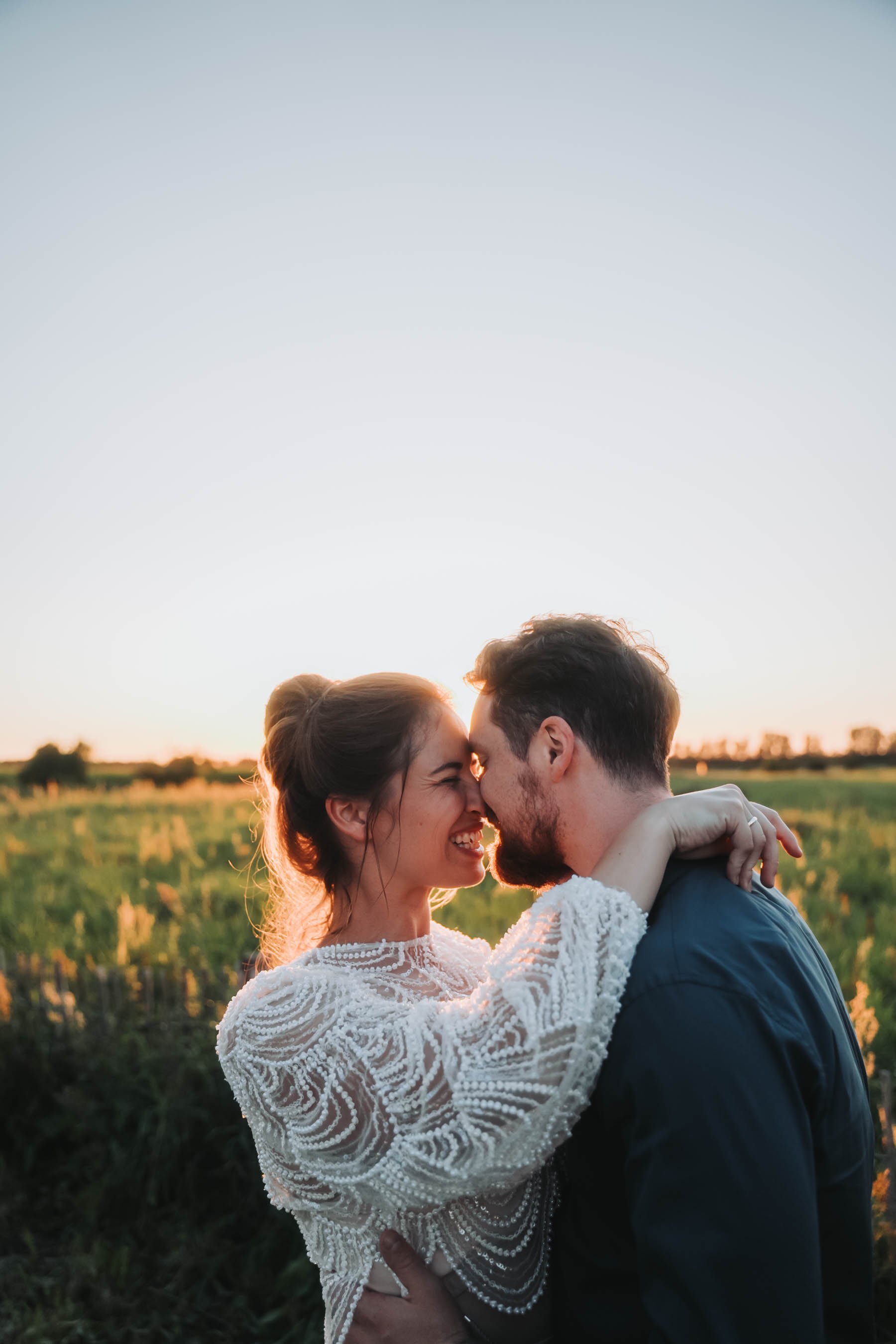 A couple embracing outdoors during sunset, with the woman wearing a white lace dress and the man in a dark suit, their foreheads touching and smiling.