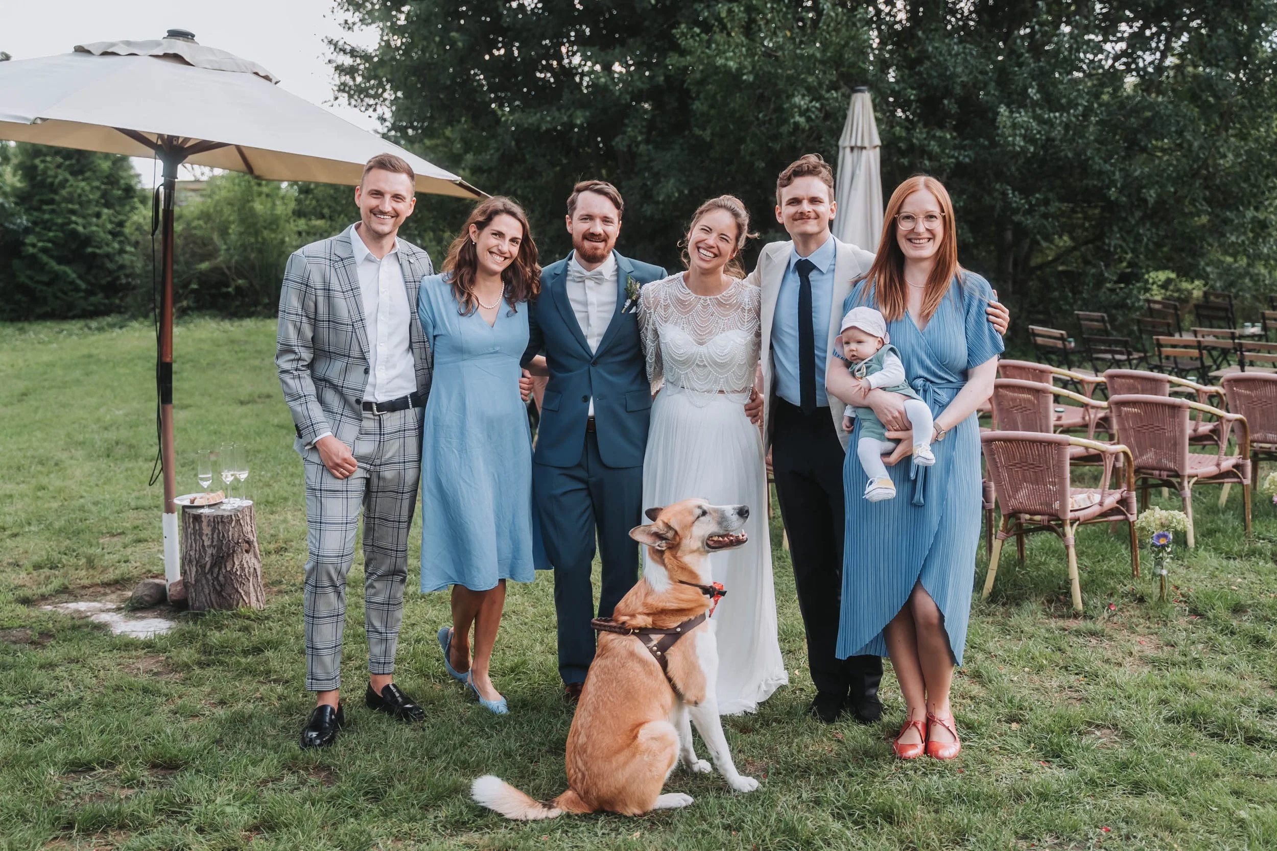 A group of seven people, including two children, smiling and standing outdoors at a wedding with trees and chairs in the background. Two dogs are also present, one sitting and one standing. The bride and groom are in the center, with friends and fami