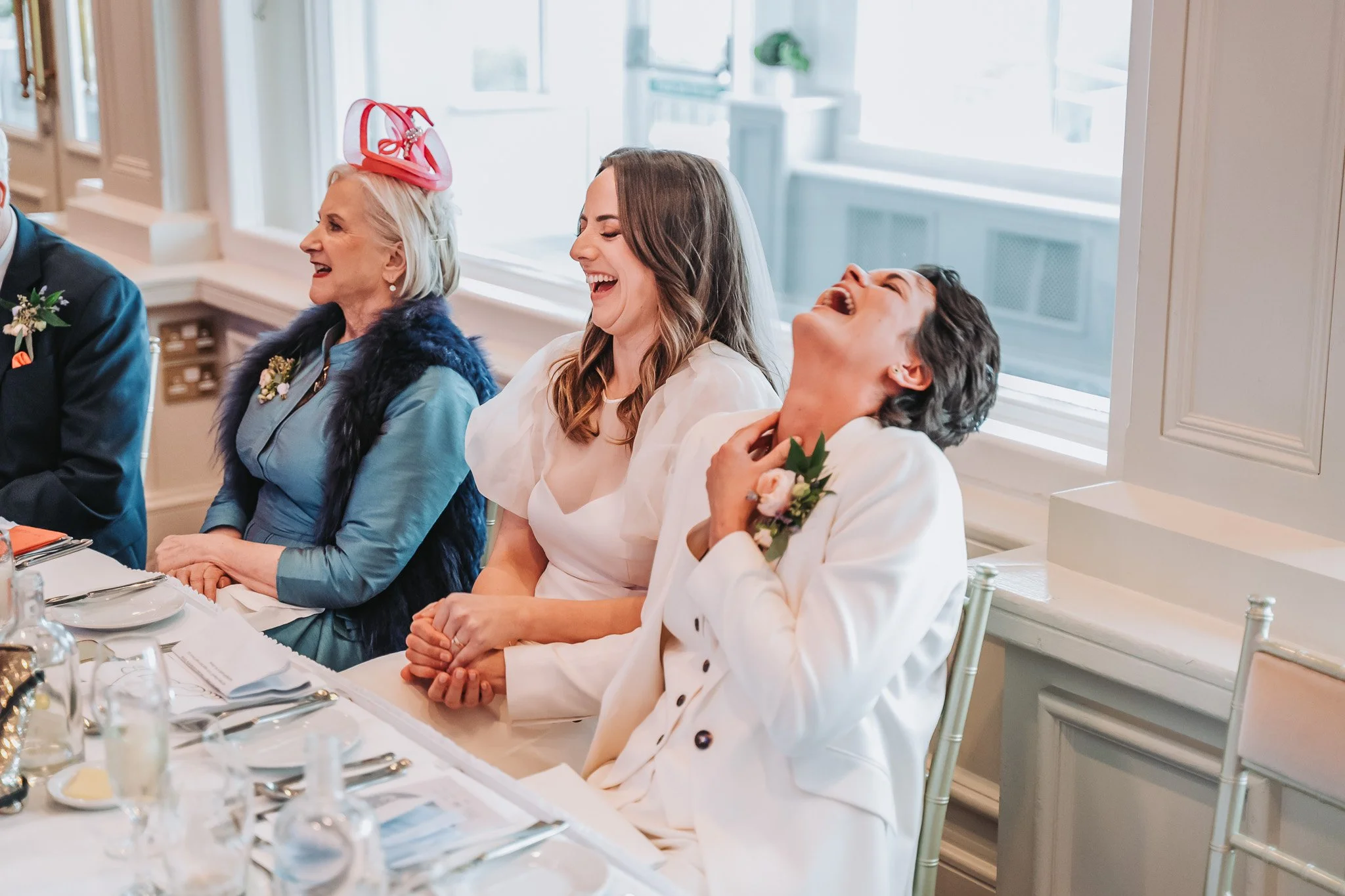 A group of women and a man sitting at a long table, laughing and enjoying a celebration or special occasion in a well-lit room with large windows.