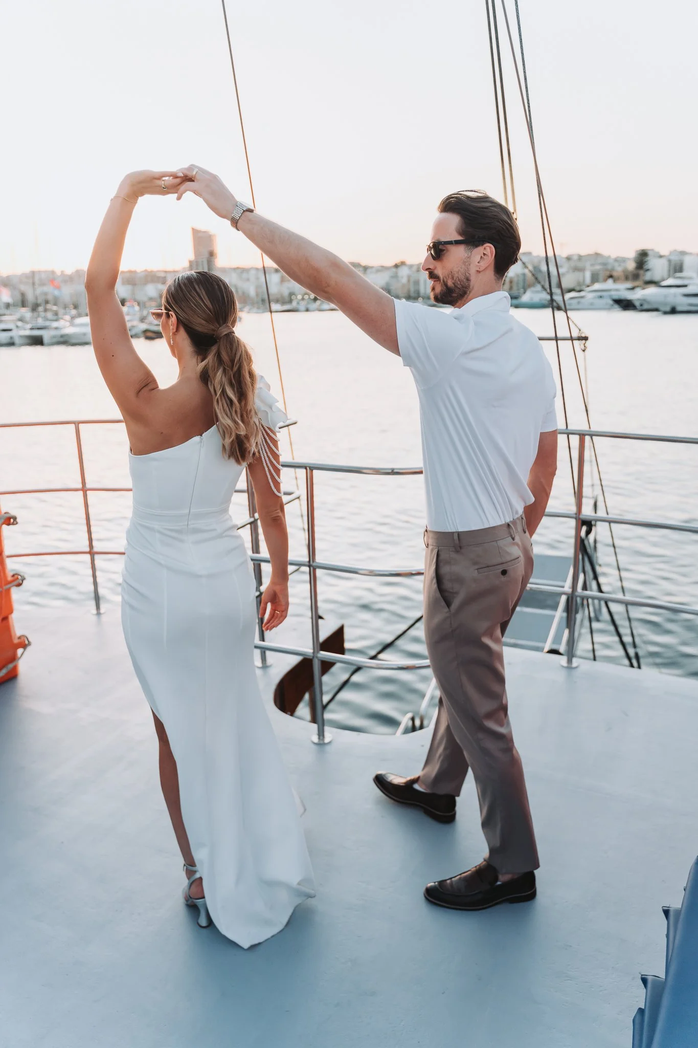 A couple dancing on a boat with a marina in the background during sunset, with the woman wearing a white dress and the man in a white shirt and gray pants.