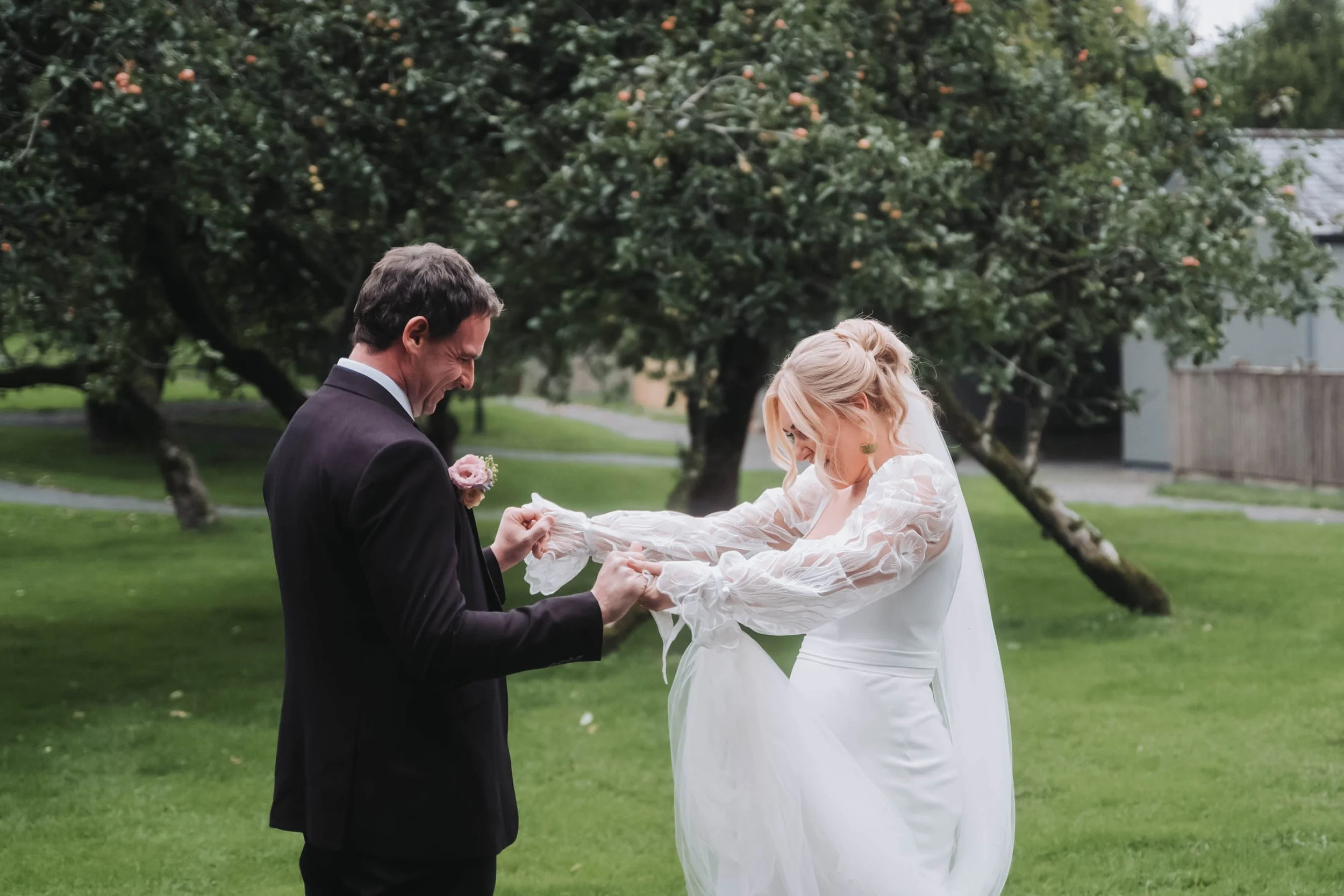 Two people, a man and a woman, are participating in a wedding ceremony outdoors. The man, wearing a black suit, is holding the woman's hands and smiling, while the woman, dressed in a white wedding gown, is looking down as they exchange vows. There a