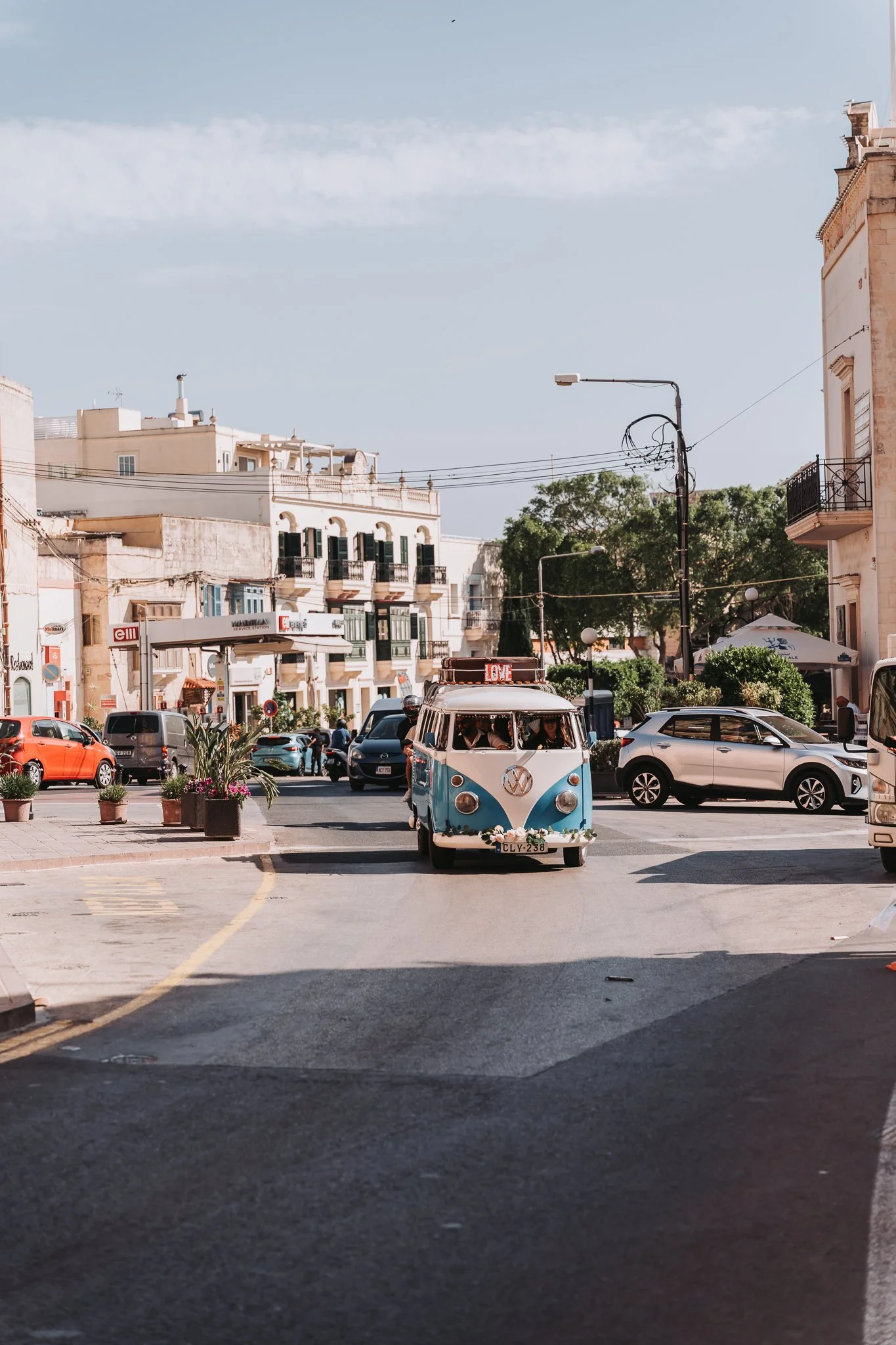 A small vintage blue and white Volkswagen van with a sign that says 'LOVE' on the roof, parked at a busy city street with cars and buildings in the background.