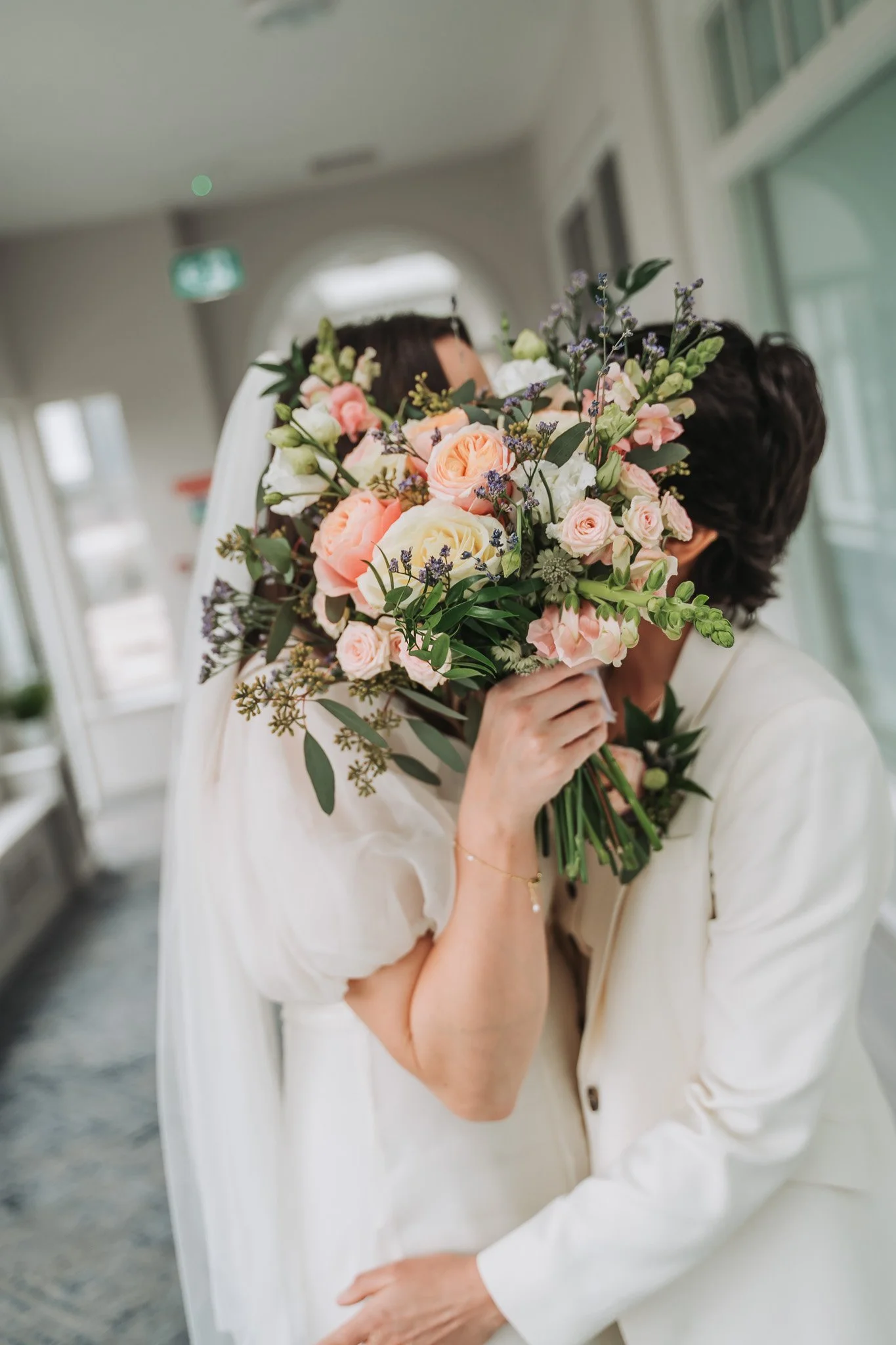 A person holding a bouquet of pink, white, and purple flowers close to their face, partially obscuring their face. The person is dressed in a light-colored outfit and is in an indoor setting with large windows and soft natural lighting.