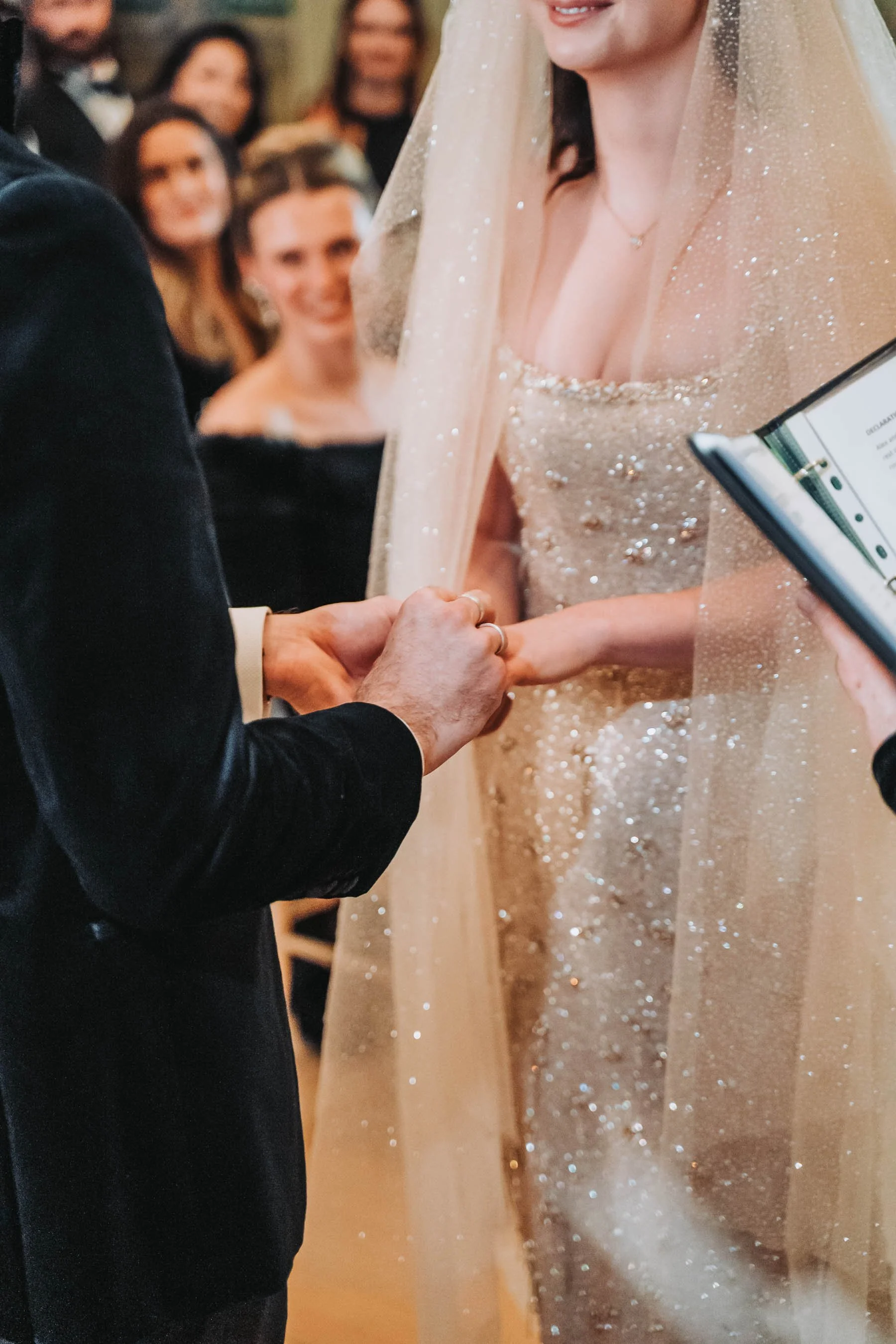 Close-up of a wedding ceremony with the bride and groom holding hands. The bride is wearing a sparkly, beige wedding gown with a veil, and the groom is in a black tuxedo. Blurred guests are seen in the background, smiling and watching the couple.