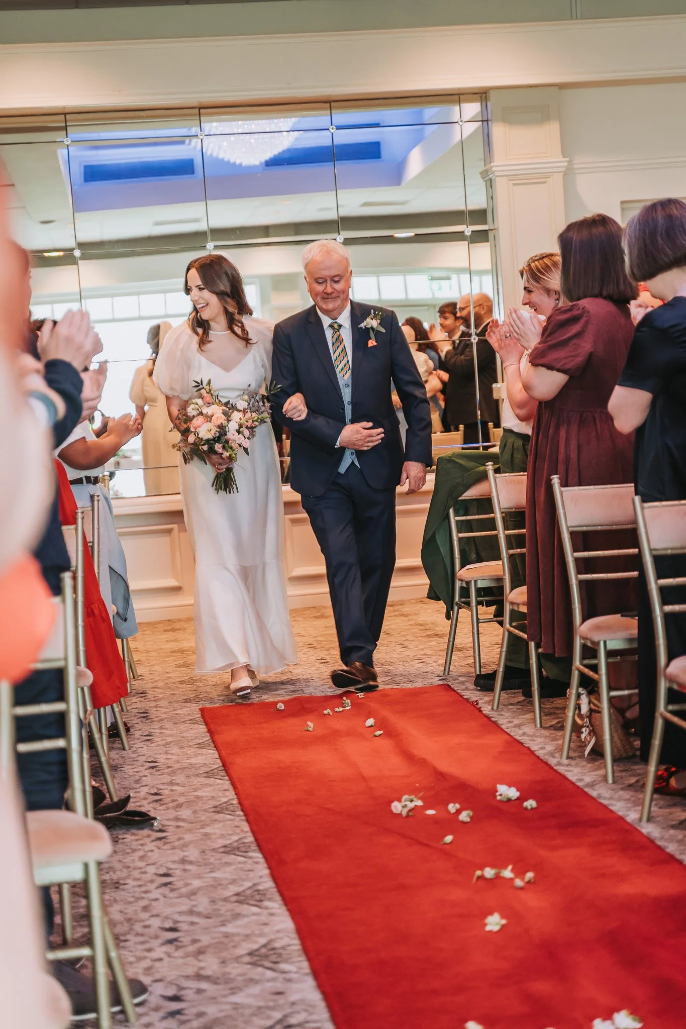 A bride walking down the aisle with her father at a wedding ceremony, surrounded by seated guests who are clapping and smiling.