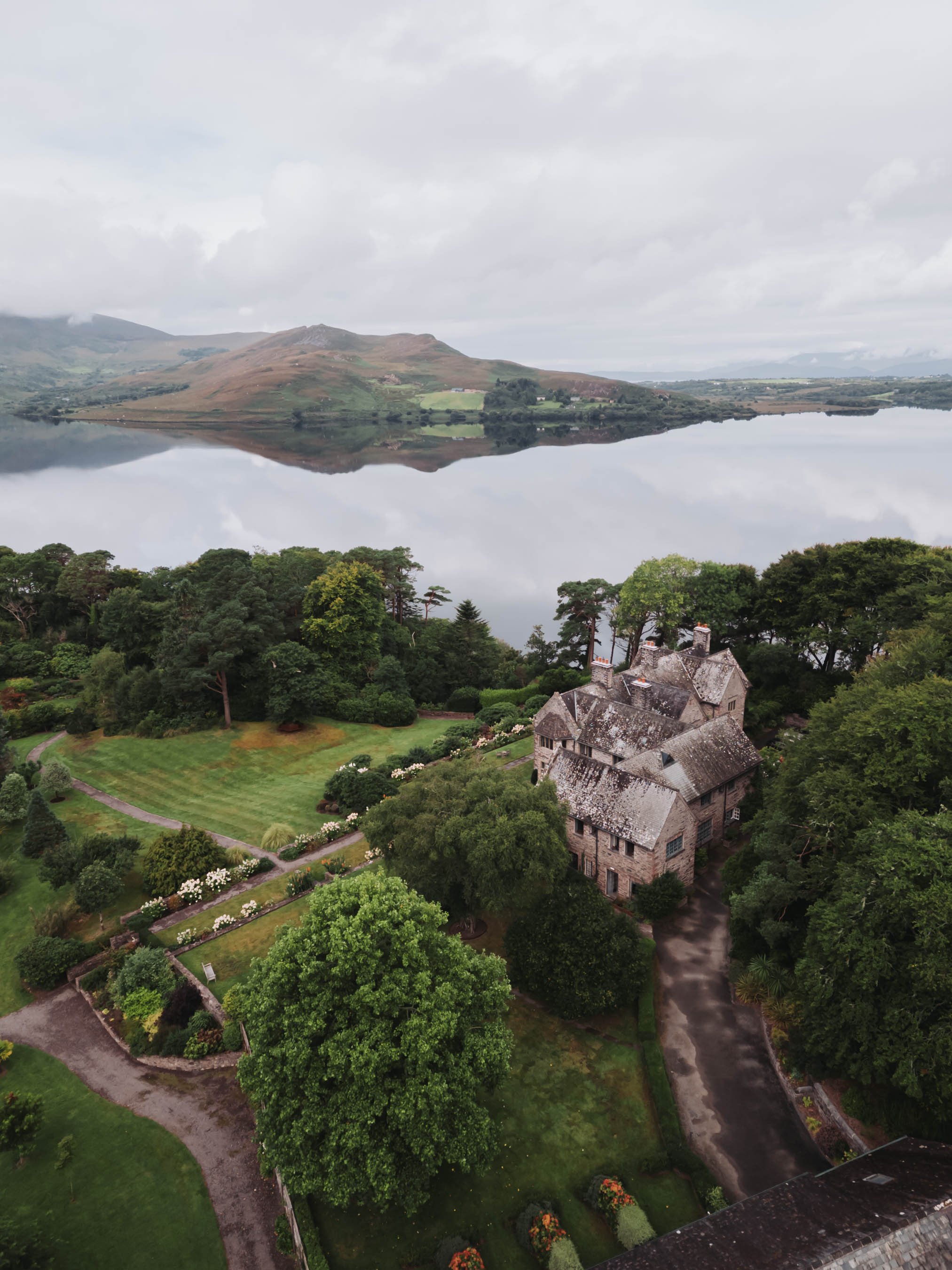 Aerial view of a historic stone mansion surrounded by lush green gardens and trees, with a calm lake reflecting rolling hills and mountains in the background under a cloudy sky.