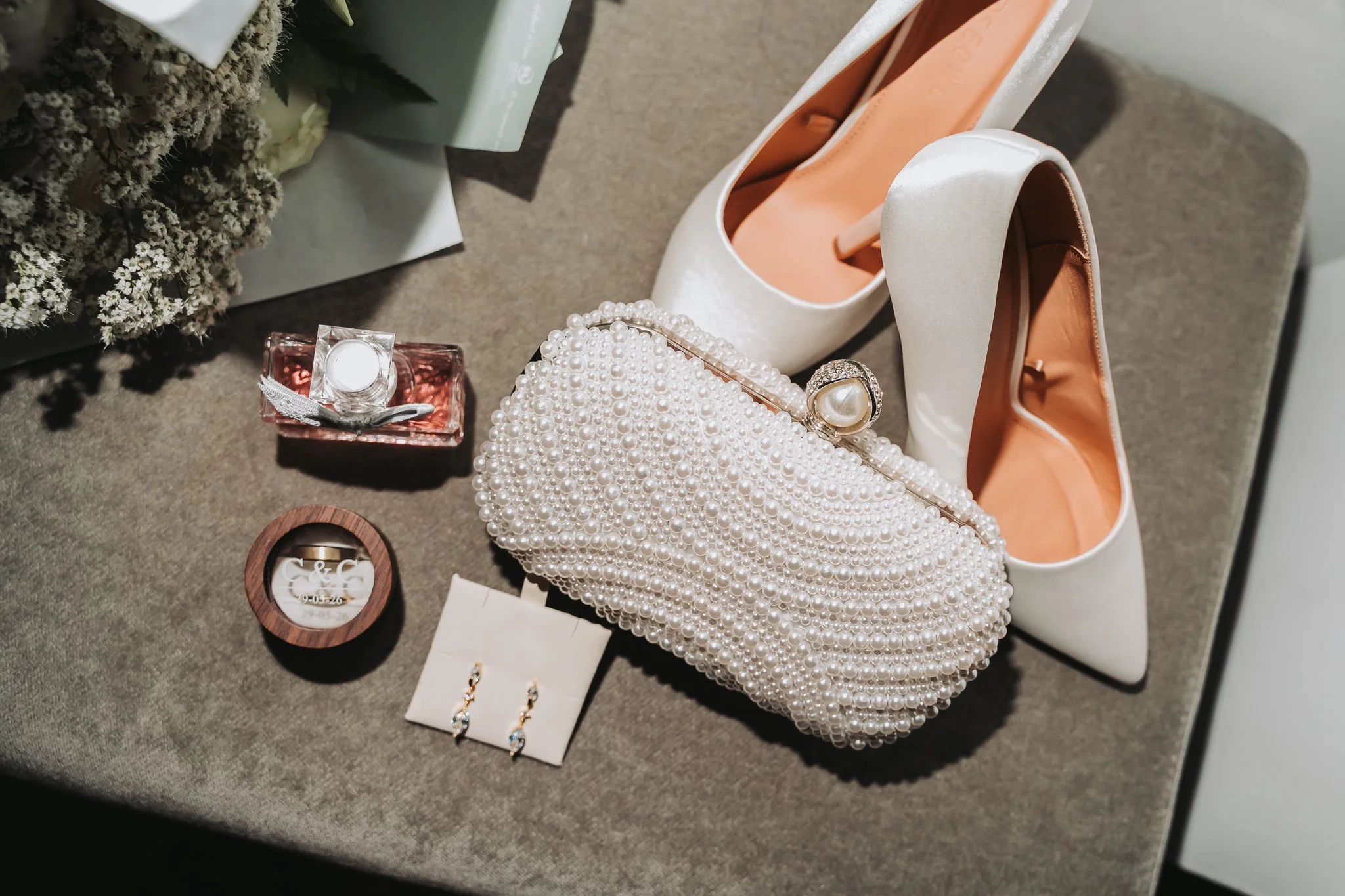 Various accessories including a pair of white shoes with peach interior, a pearl clutch, pearl earrings, a pearl ring, a ring with a large pearl and rhinestones, a small gift box with a ring, and a floral arrangement on a gray surface.