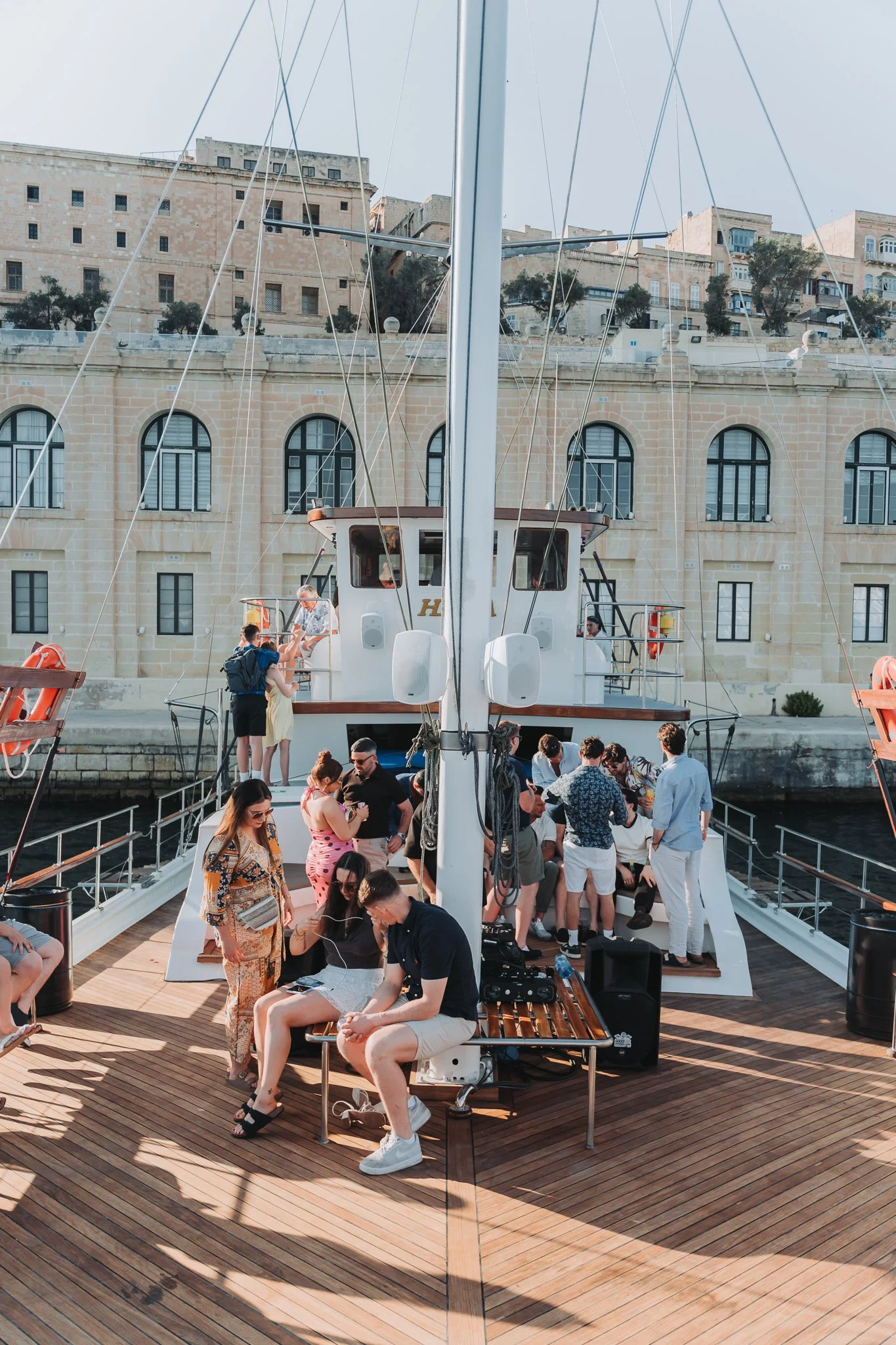 People on a boat deck, some seated and some standing, with a large ship in the background and historic buildings beyond, during daytime.