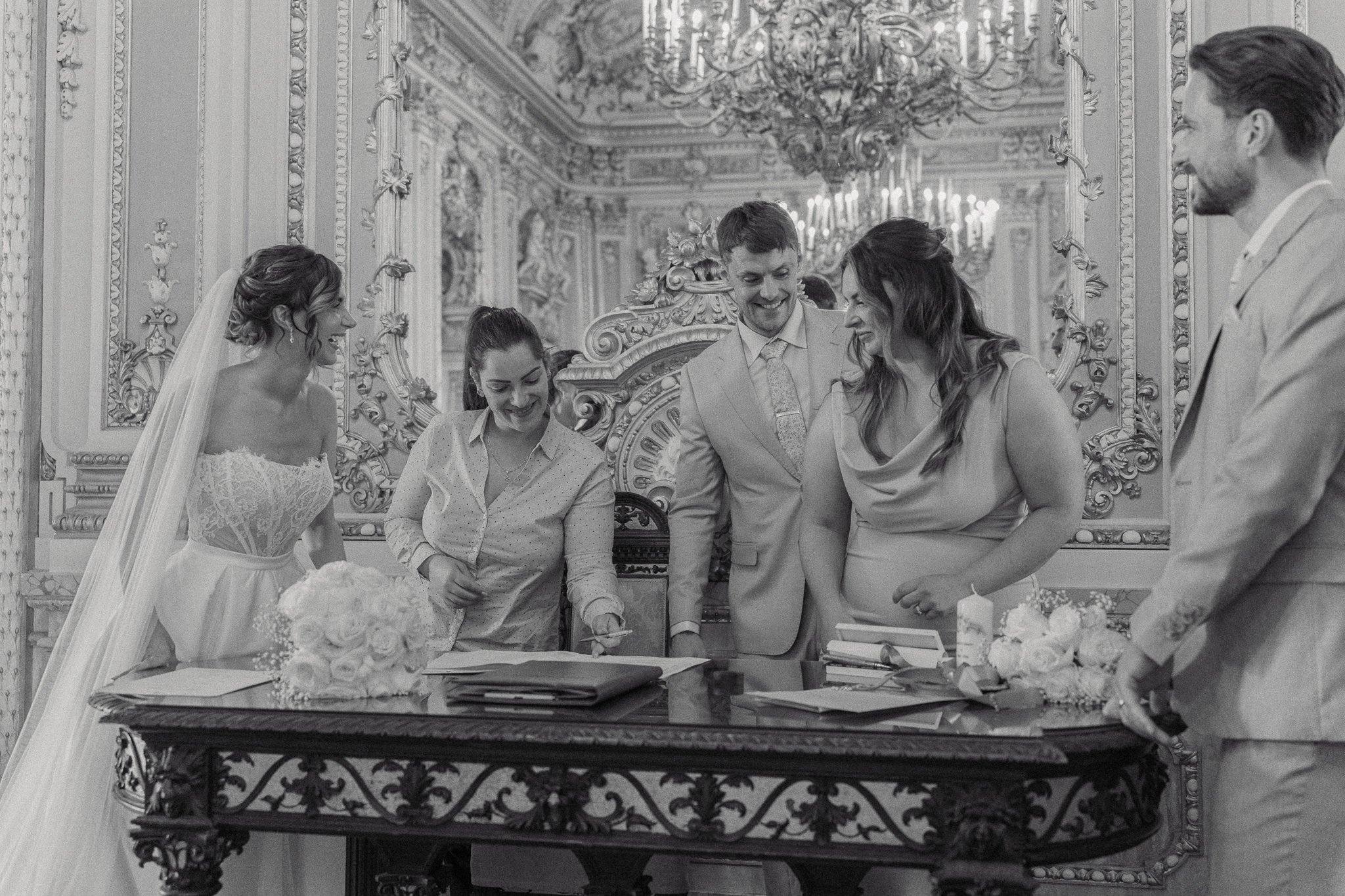 A group of five people, two women and three men, gathered around a table, signing documents during a wedding ceremony in an ornate room with a chandelier and detailed wall moldings.