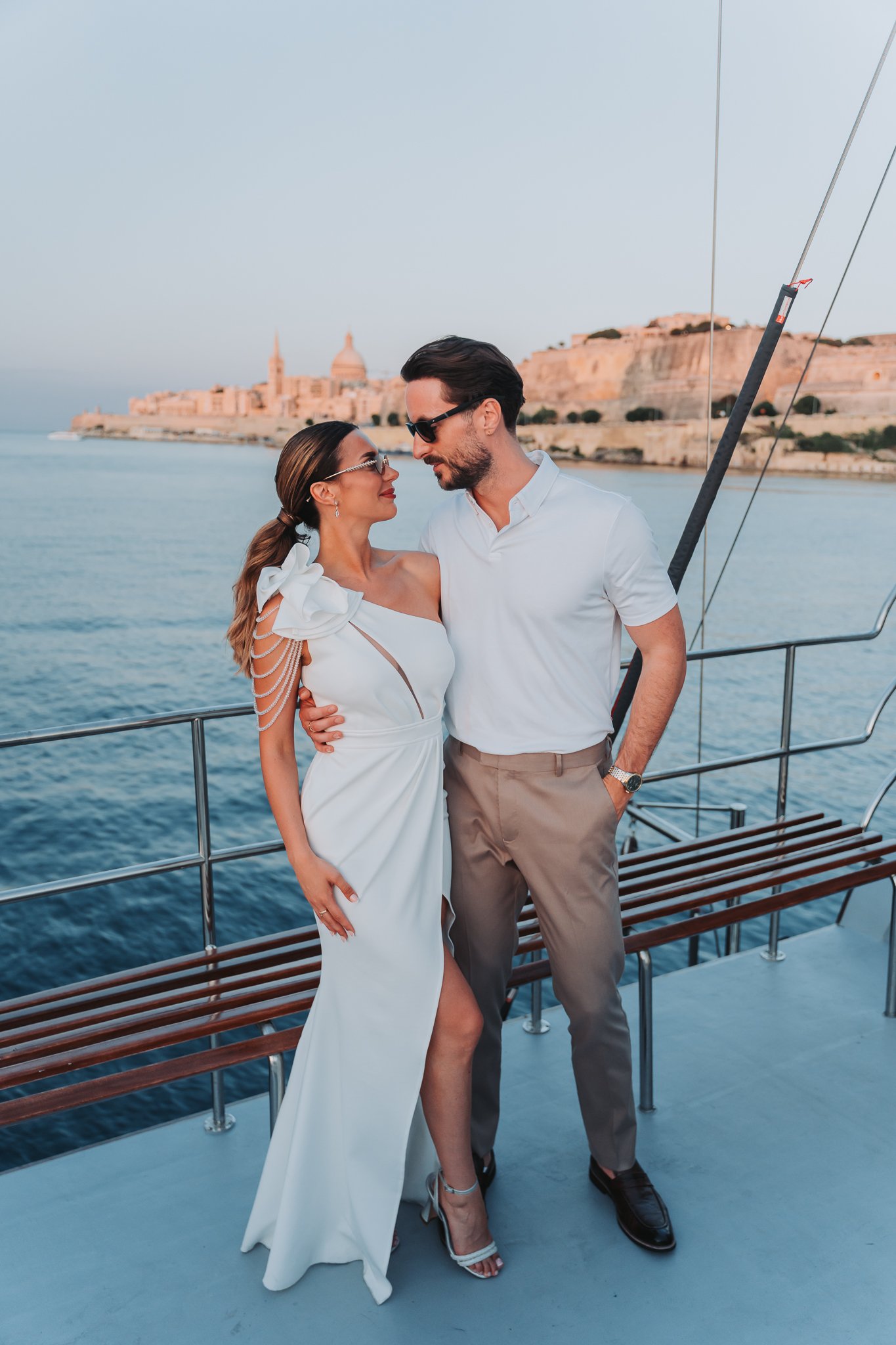A couple standing on a boat deck near water, with a historic cityscape in the background, including domed buildings and church steeples, during sunset.