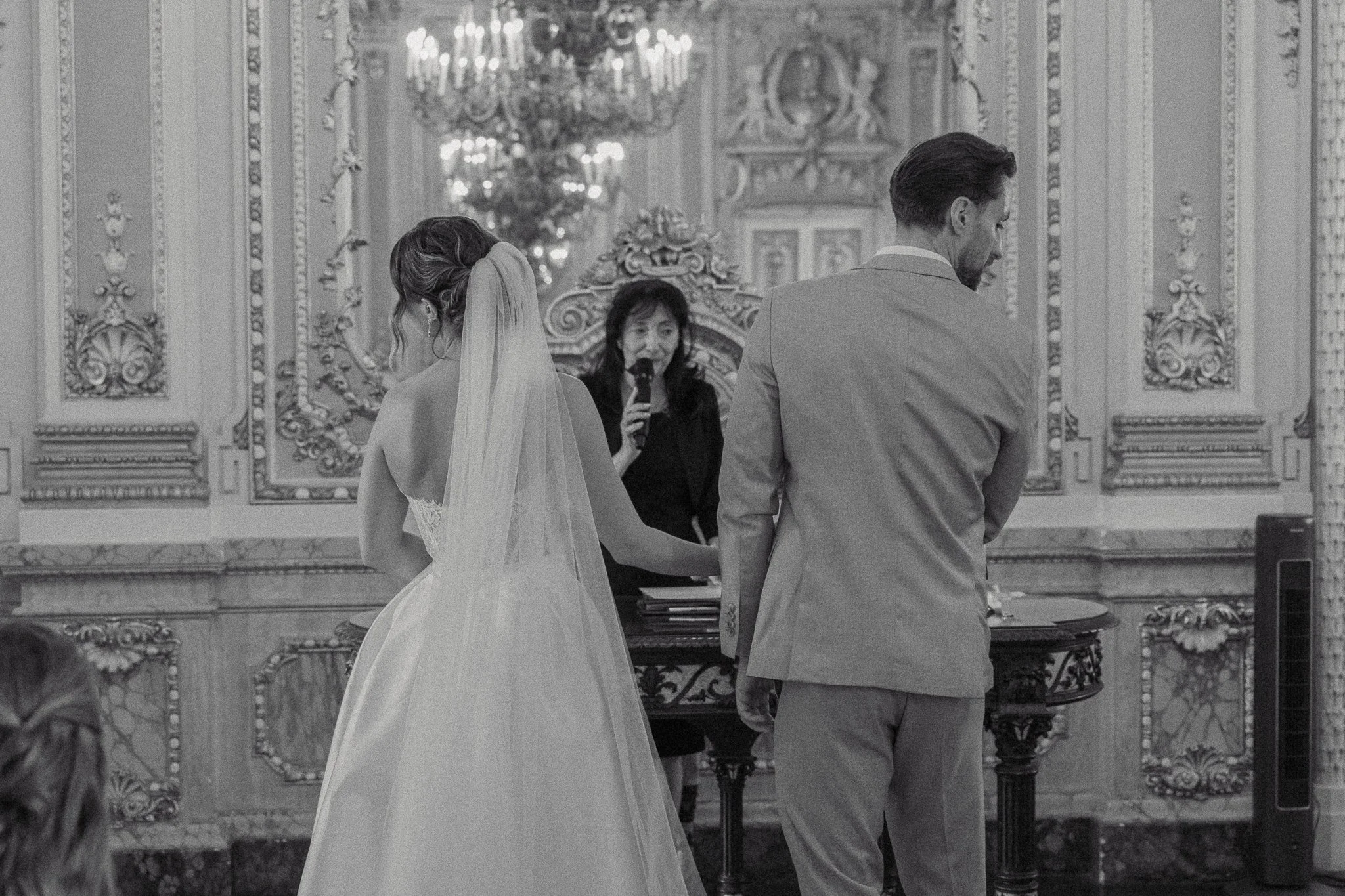 A bride and groom standing with an officiant during a wedding ceremony in an ornate, historic hall with detailed wall decorations and a chandelier.