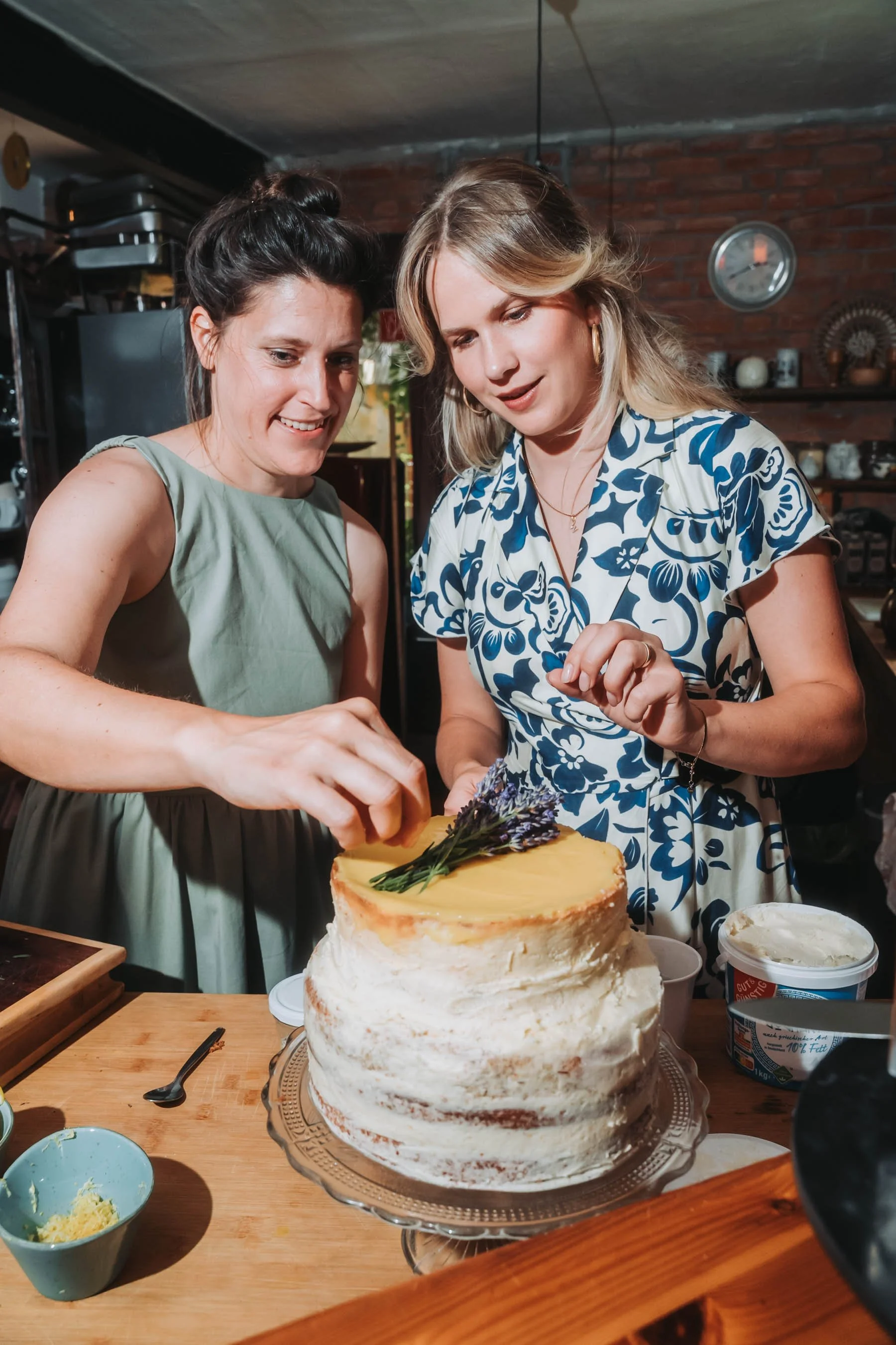 Two women standing behind a layered cake on a cake stand, decorating it with herbs and flowers. The cake has white icing and is placed on a wooden table with utensils and ingredients around.