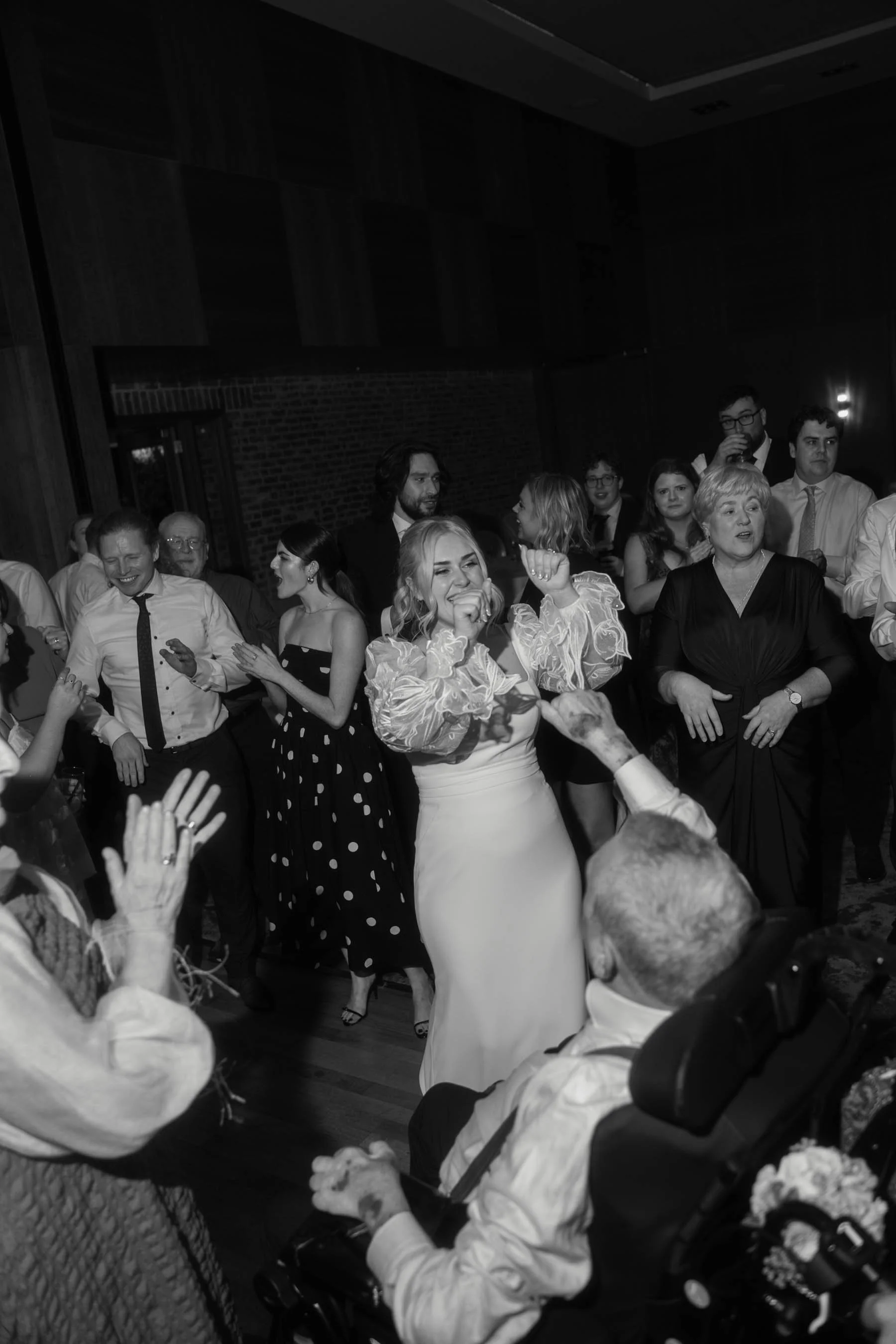 A group of people dancing and celebrating at an indoor party or event, with a woman in a white dress and other guests in formal attire.