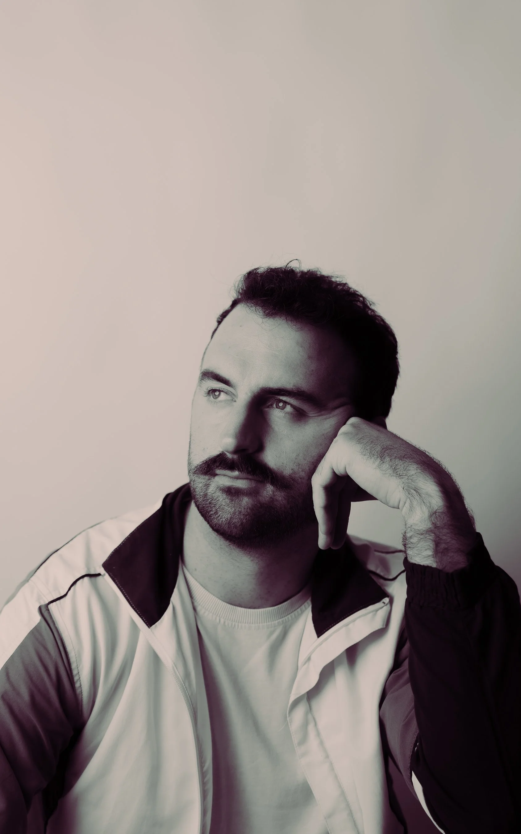 A black and white portrait of a man with dark hair, a mustache, and a beard, wearing a sports jacket, resting his head on his hand while looking thoughtfully to the side.