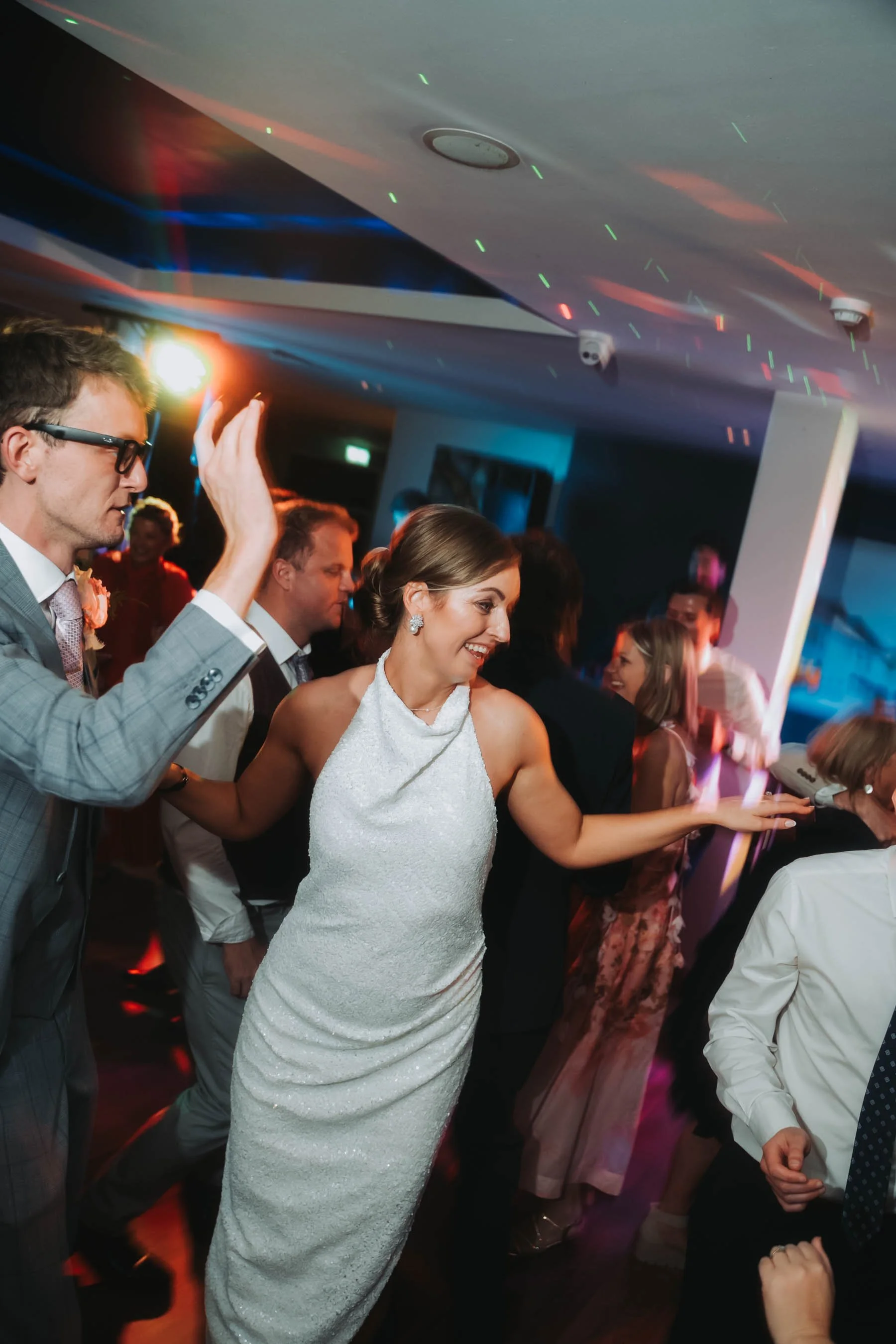 People dancing at a celebration or wedding reception with colorful lighting and a lively atmosphere.