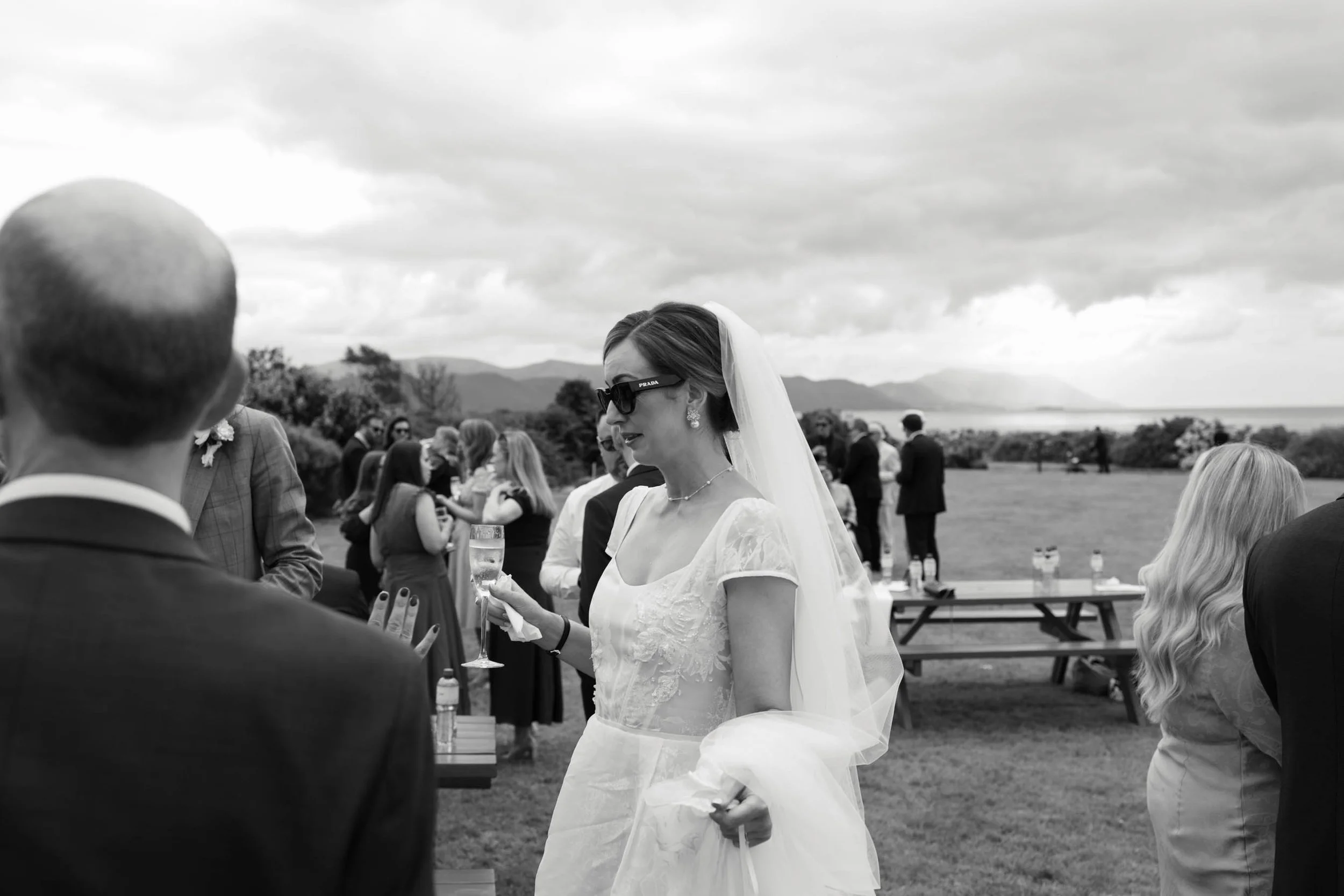 Black and white photo of a bride holding a glass at an outdoor wedding ceremony, with guests in the background and mountains in the distance.
