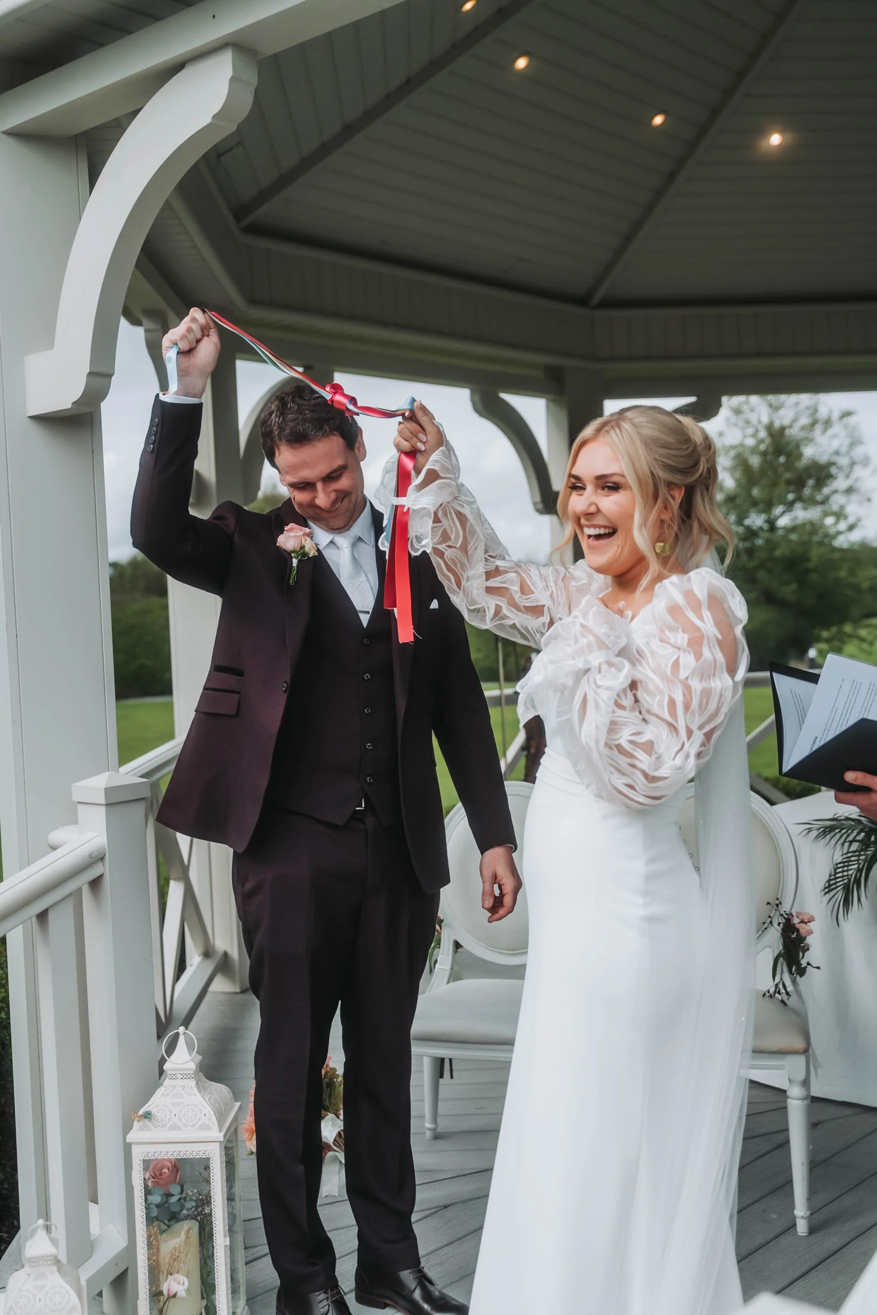 A bride and groom celebrating their wedding outdoors on a decorated porch, with the bride smiling and holding a ribbon that loops around the groom's neck.