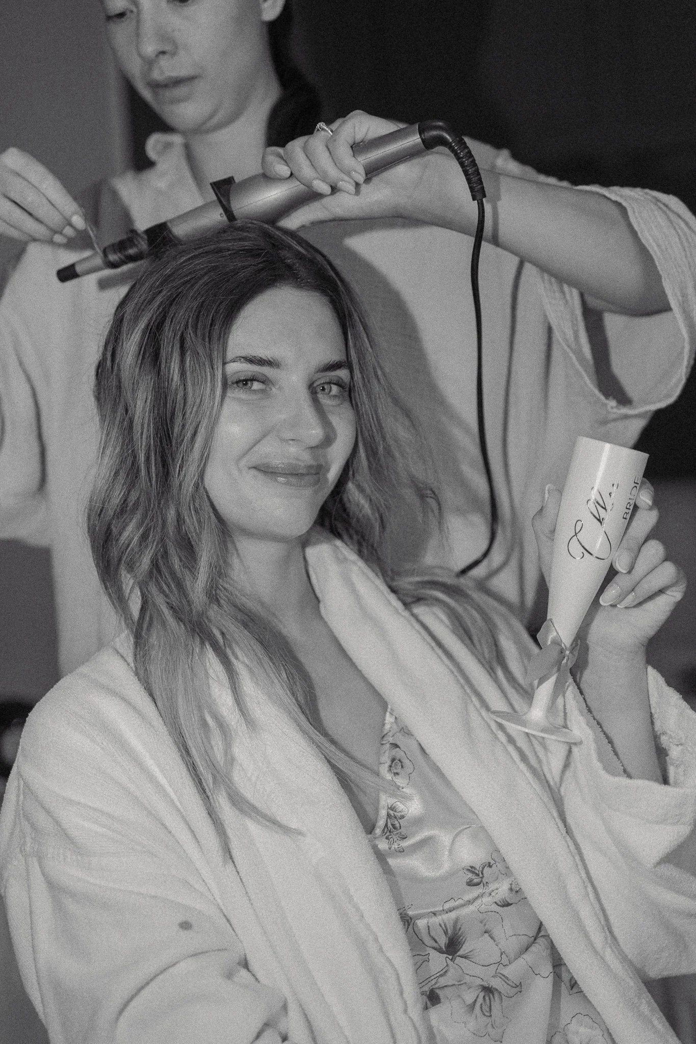 A woman with long hair smiling as she holds a bouquet of flowers and a white cup, while a hair styling device is being used on her hair by another person.