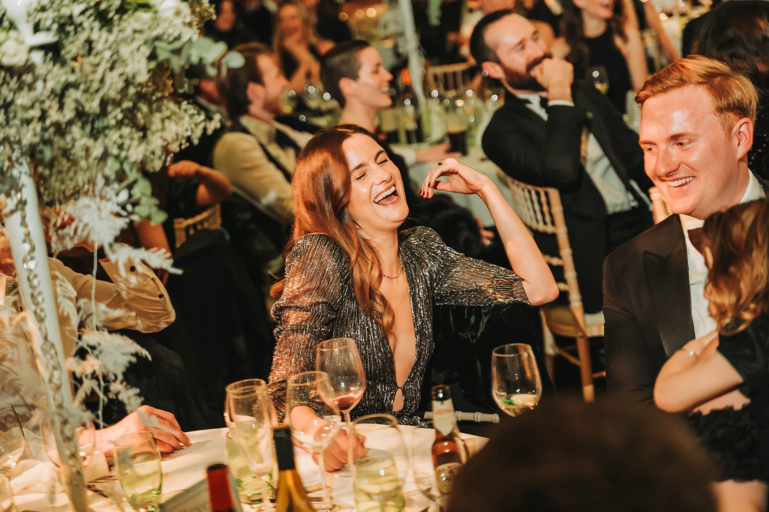 People attending a formal event or celebration, sitting at a decorated table with drinks, dressed in elegant clothing, smiling and laughing.
