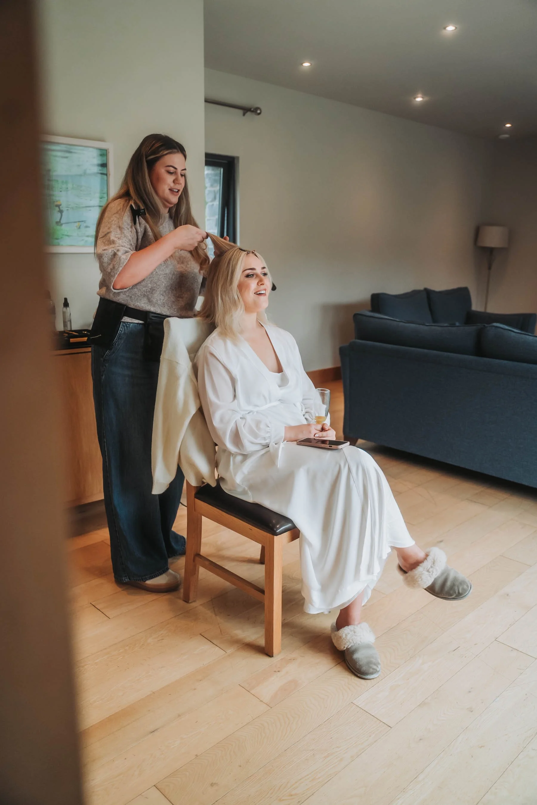 A woman sits on a wooden chair wearing a white robe and fluffy slippers, holding a smartphone with a glass of wine in her lap, while a hairstylist styles her blonde hair in a cozy, well-lit living room.