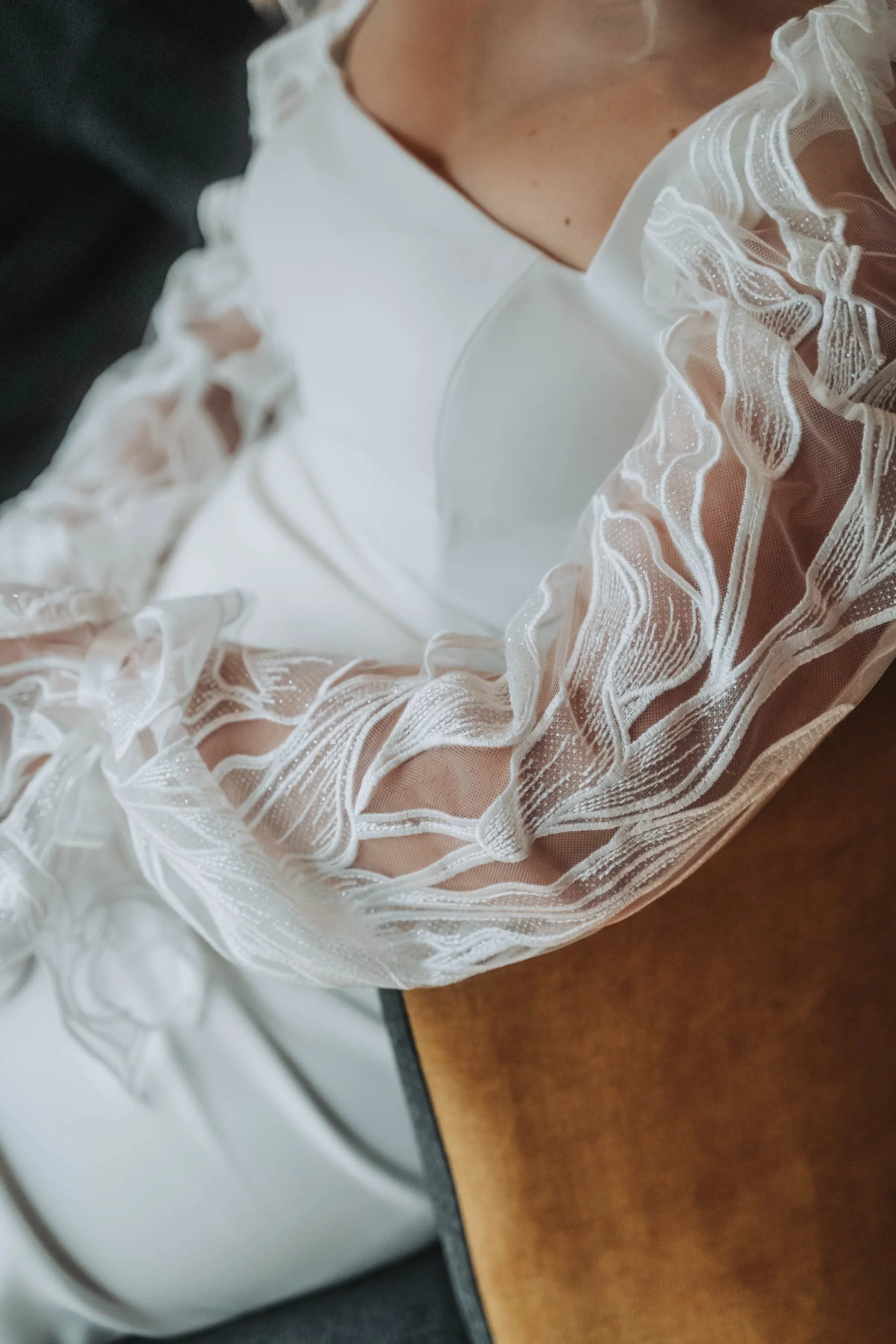 Close-up of a person's chest wearing a white garment with sheer, ruffled lace sleeves featuring intricate embroidered and sparkly details.