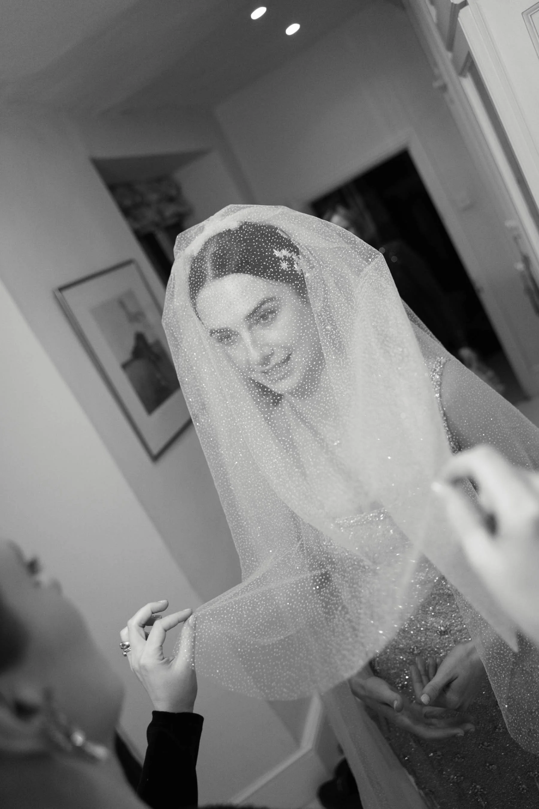 A woman with dark hair and makeup wears a sparkling, sheer veil with a snowflake pattern, smiling and looking at her reflection in a mirror. A person in a black long sleeve shirt is adjusting or touching the veil. The background shows a room with fra