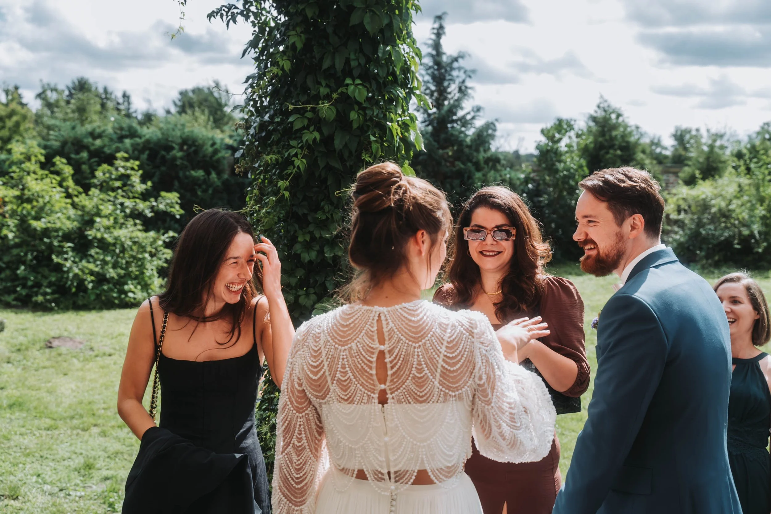 Group of people, including women and a man, gathered outdoors in front of greenery, smiling and engaging in conversation, with one woman wearing glasses and a wedding dress.