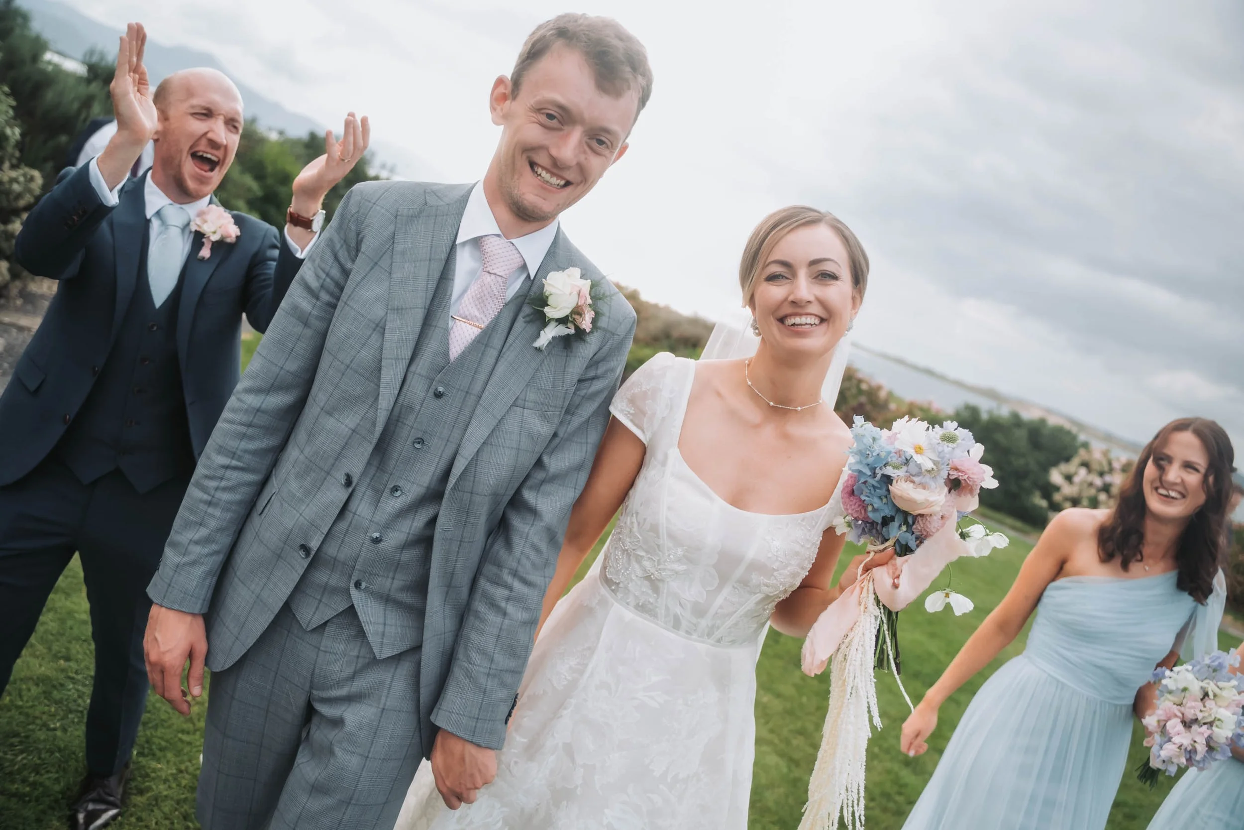 Bride and groom smiling and holding hands at an outdoor wedding ceremony, surrounded by bridesmaids and a groomsman. The groom is in a gray checked suit and pink tie, and the bride wears a white lace wedding dress, holding a bouquet of flowers. The g