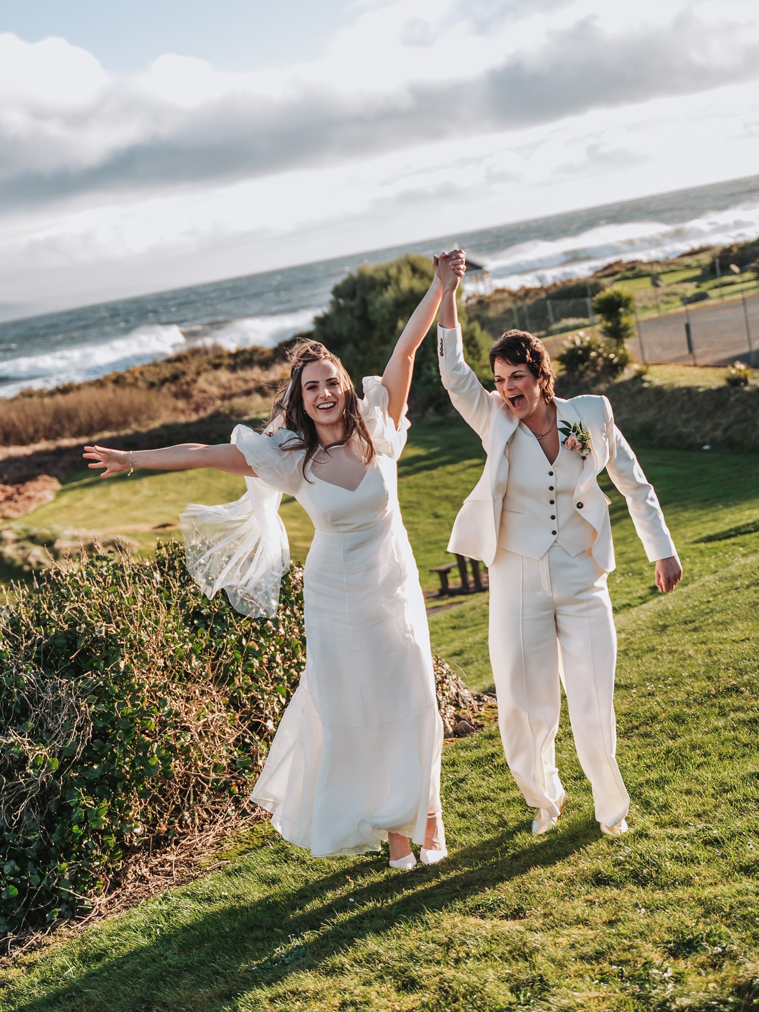 Two women in white wedding dresses holding hands up in celebration on a grassy outdoor area near the ocean, with cloudy sky overhead.