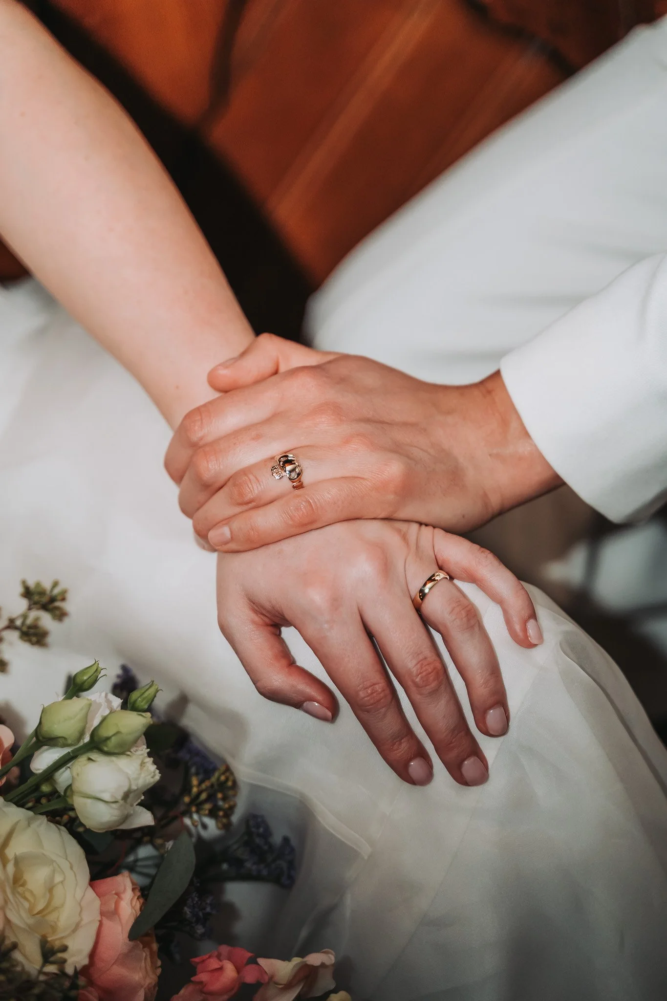 Close-up of two hands gently holding each other, with one hand wearing a gold ring with a diamond. The background includes white and pink roses and flowers, on a white surface.
