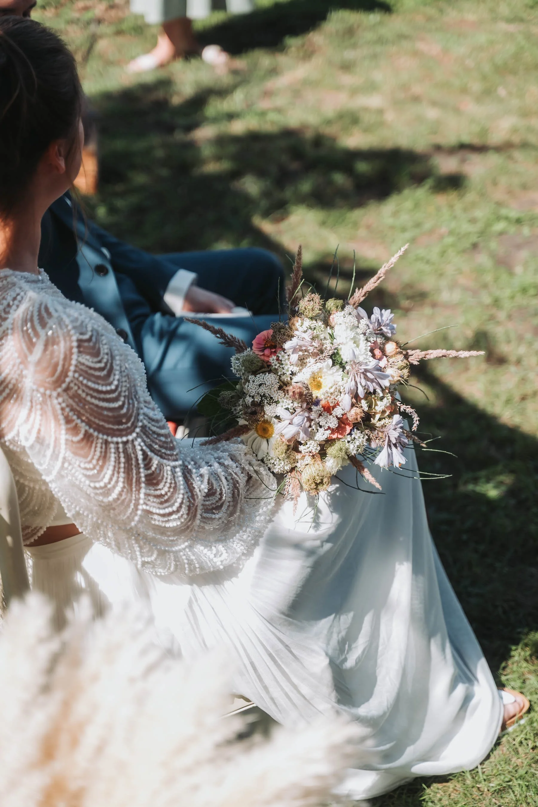 A woman in a beaded dress holds a bouquet of flowers while sitting on grass outdoors. A person in a suit is seated nearby, and the photo appears to be taken at a wedding or special event.