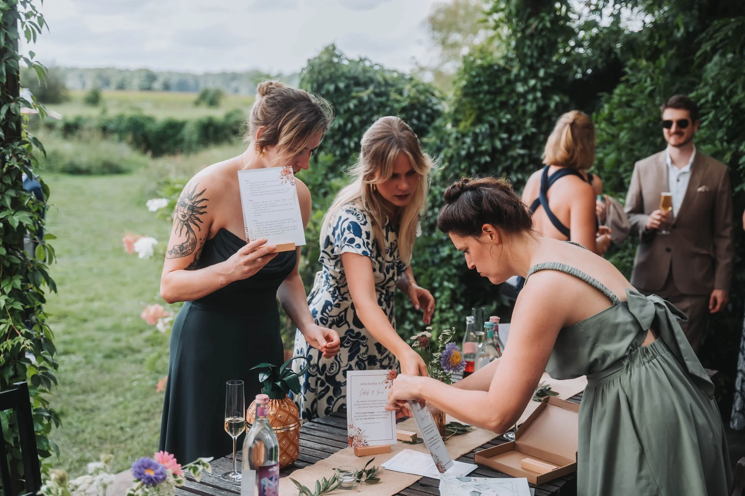 A group of women gathering outdoors around a table with flowers and drinks, with a man in the background holding a glass, on a lush green landscape.