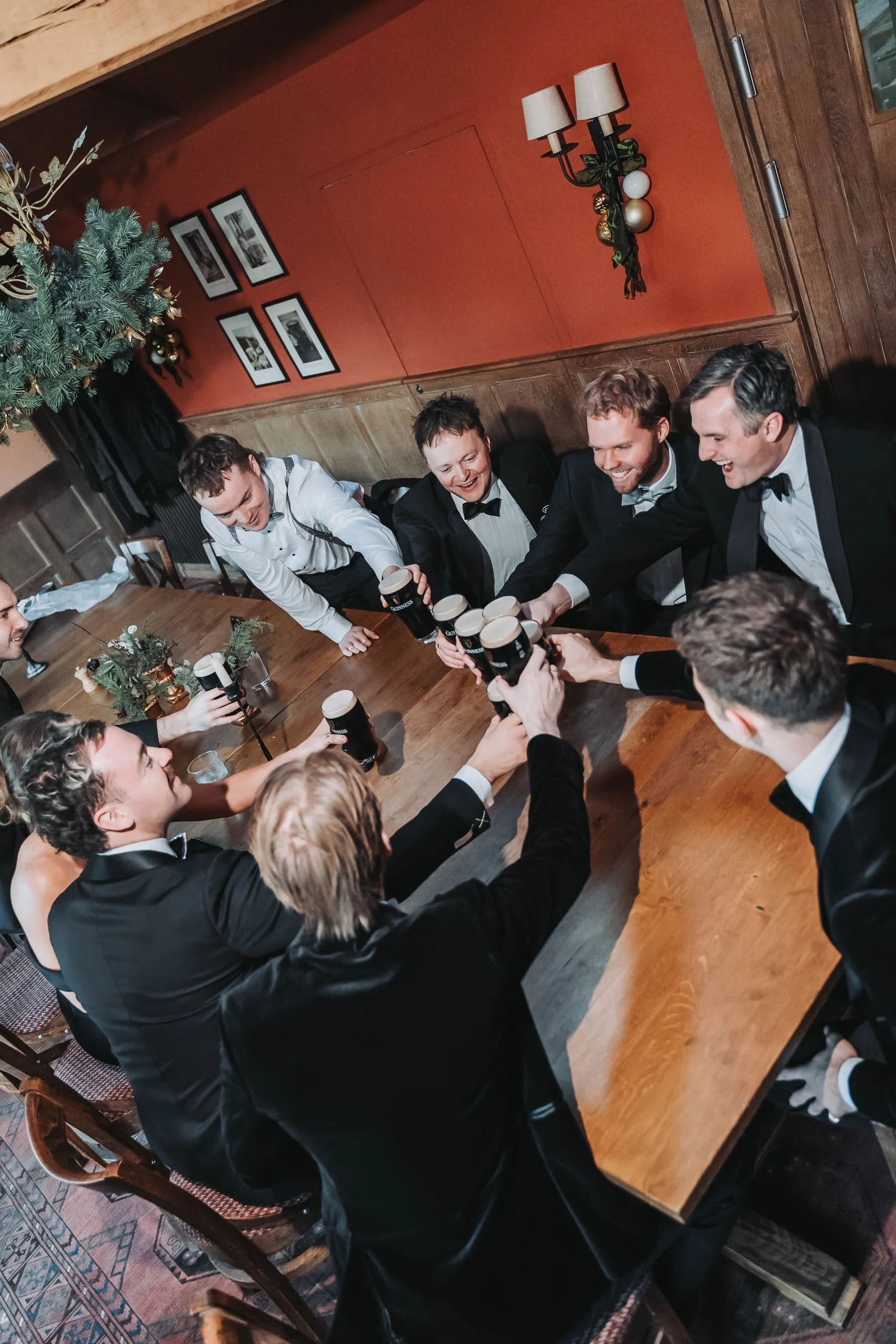A group of men in tuxedos and women in formal dresses sitting around a wooden dining table, raising pint glasses of Guinness for a toast in a decorated room with red walls, framed black and white photographs, a Christmas tree, and festive ornaments.