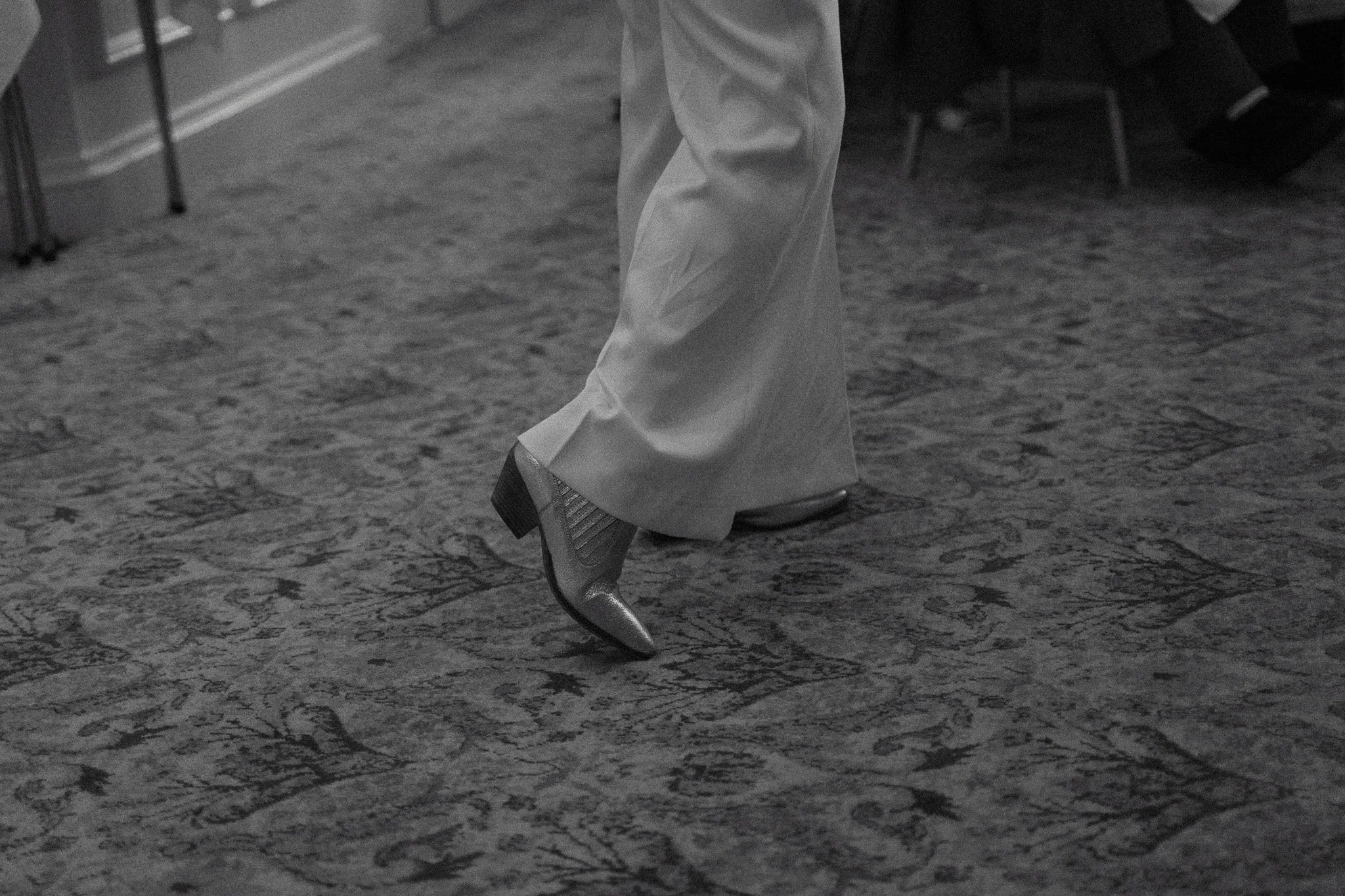 Close-up of a person's leg wearing a high-heeled shoe and wide-legged pants, standing on a patterned carpet. The setting appears to be an indoor event or gathering.