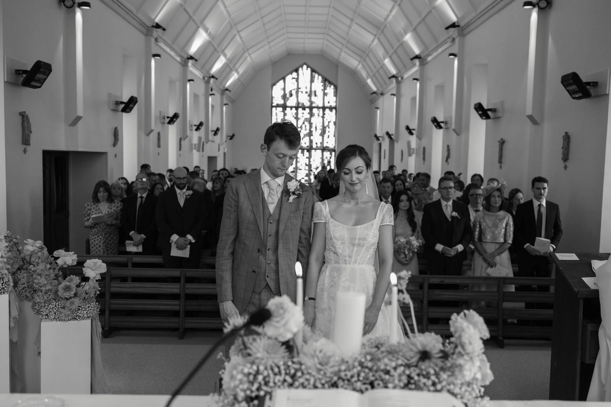 A couple standing at the altar during a wedding ceremony in a church, with guests seated behind them, black and white photo.