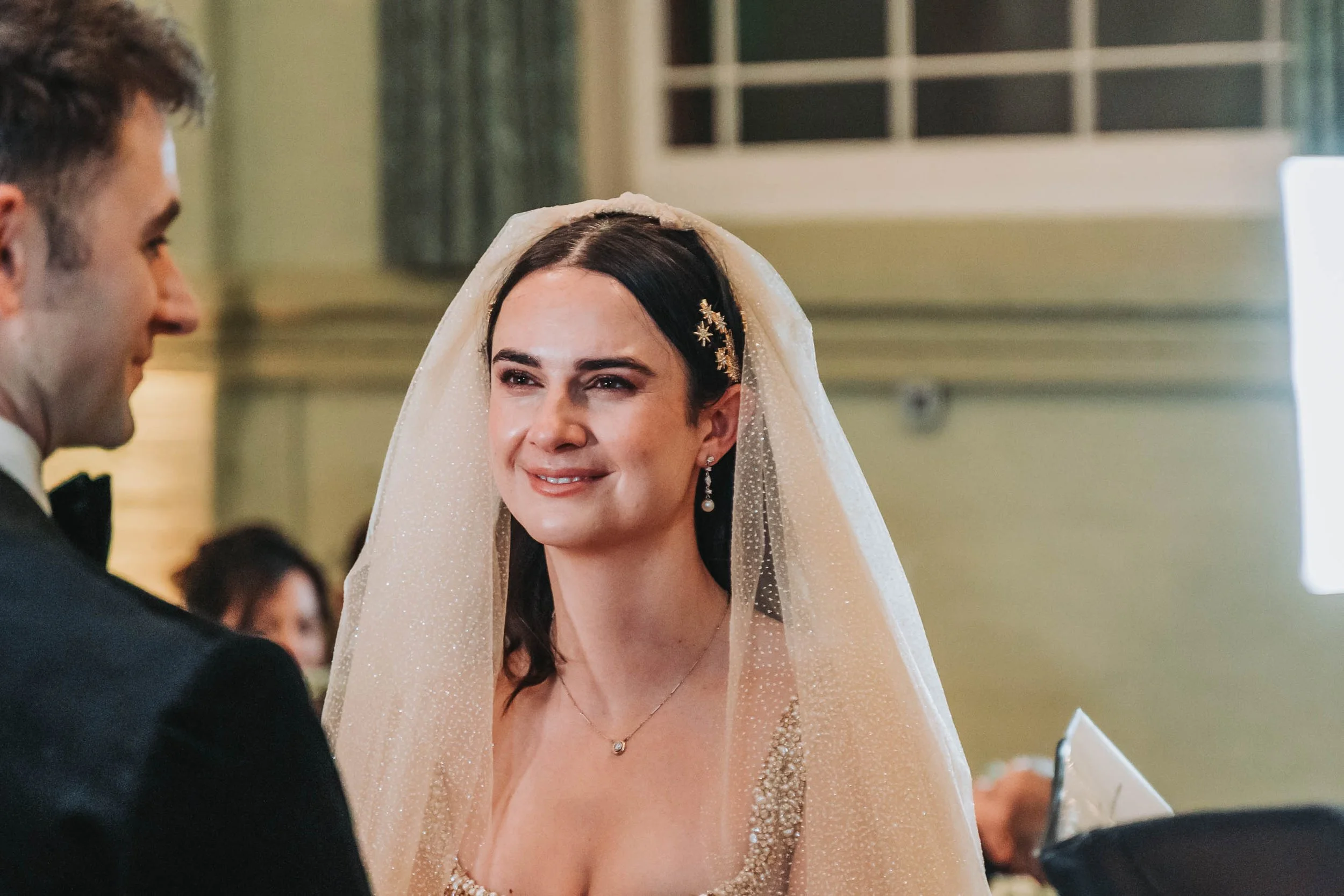 A bride with dark hair and fair skin wearing a white veil with gold star-shaped hair accessories, smiling at a groom dressed in a tuxedo during a wedding ceremony.