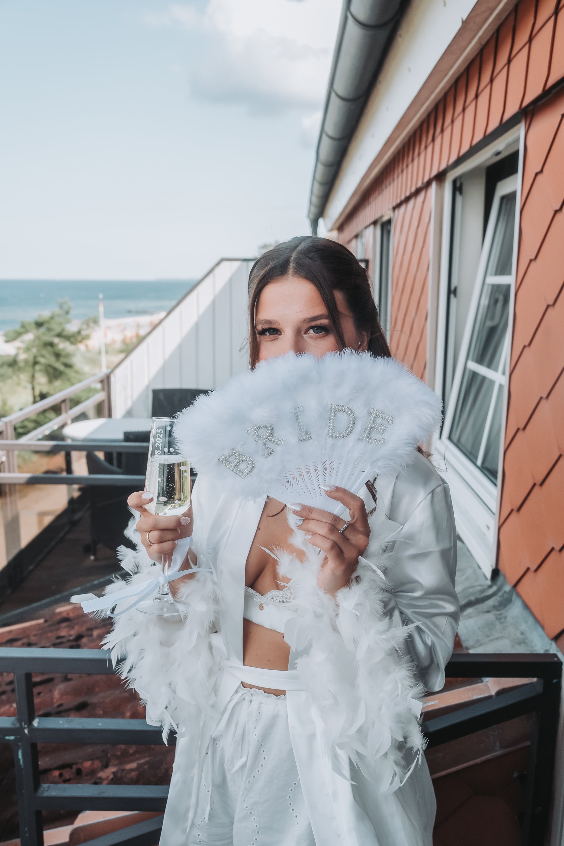 A young woman dressed in white with a feather boa and headband, holding a 'BRIDE' hand fan and a champagne glass, standing on a balcony with ocean view.