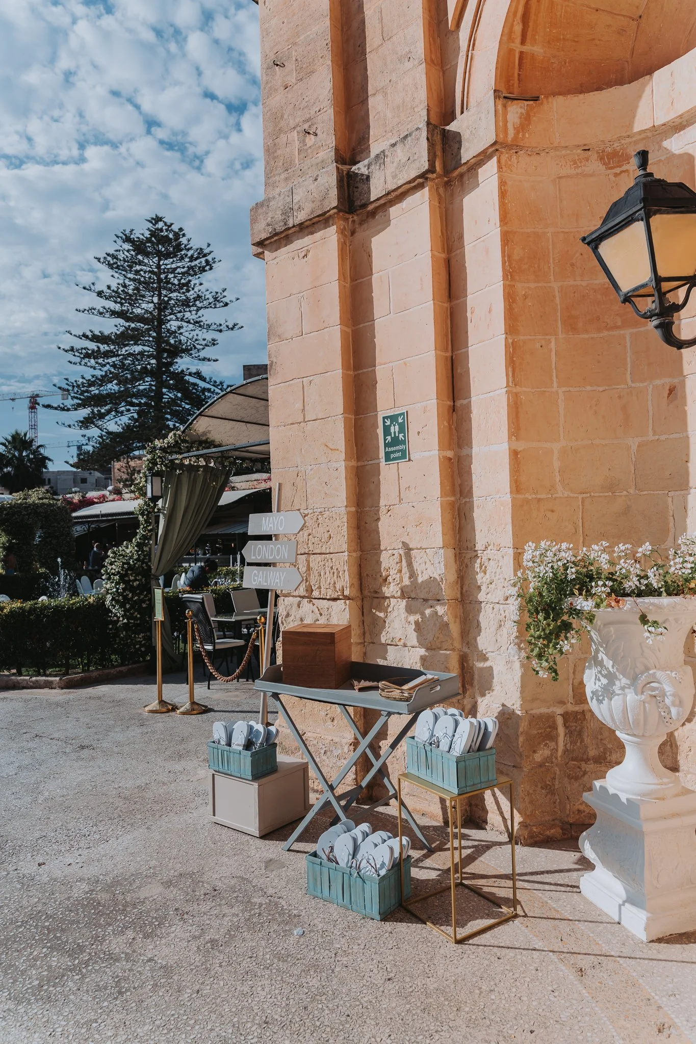 Outdoor scene with a stone building wall, flower arrangements, and a large white classical vase. There are tables with baskets of flip-flops and signs pointing to different cities.