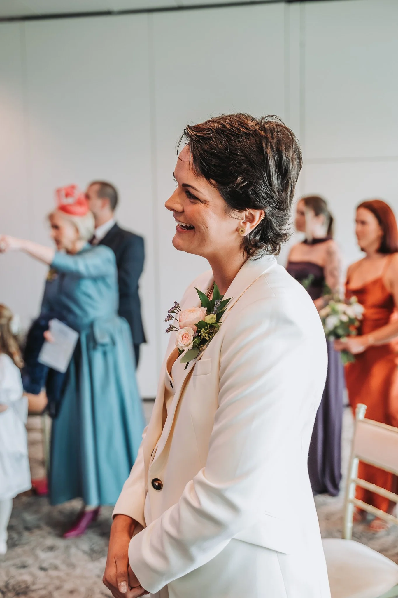 A woman with short dark hair wearing a white blazer with a floral corsage, smiling and standing among others at what appears to be a formal event or celebration in a room with white walls.