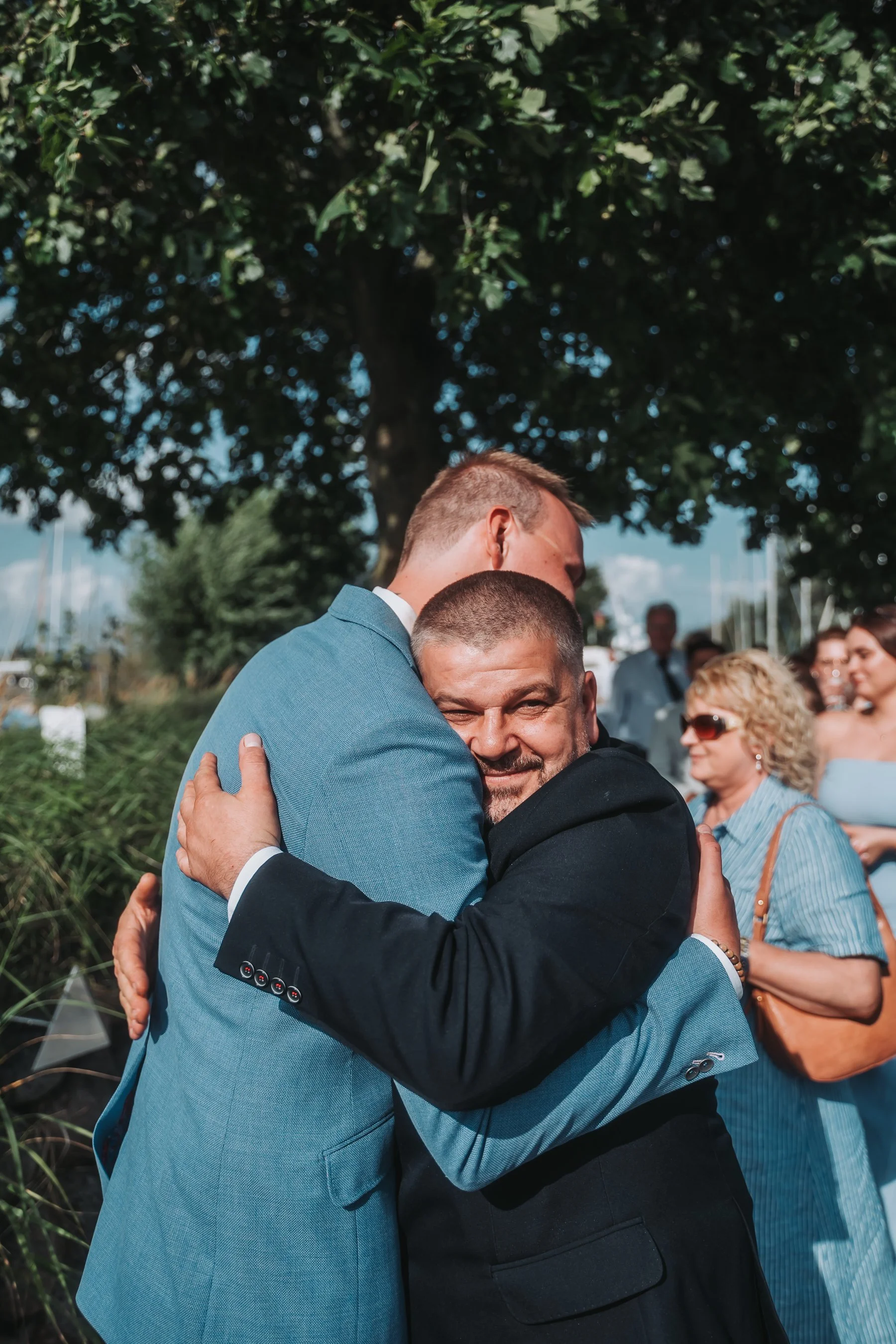 Two men in suits hugging each other outdoors at a social gathering, with a crowd of people and a large tree in the background.