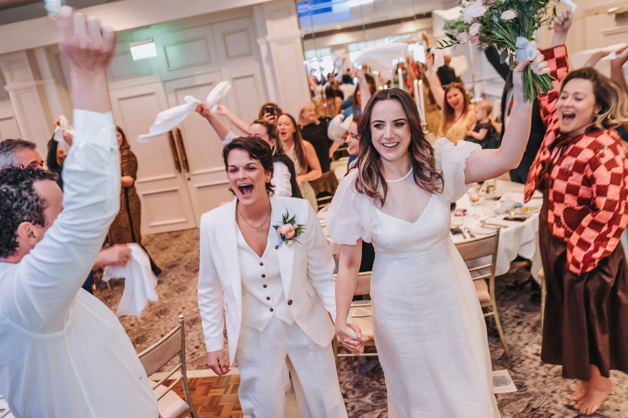 People celebrating at a wedding reception, with two women in white wedding dresses holding hands and smiling, others in the background cheering and taking photos.