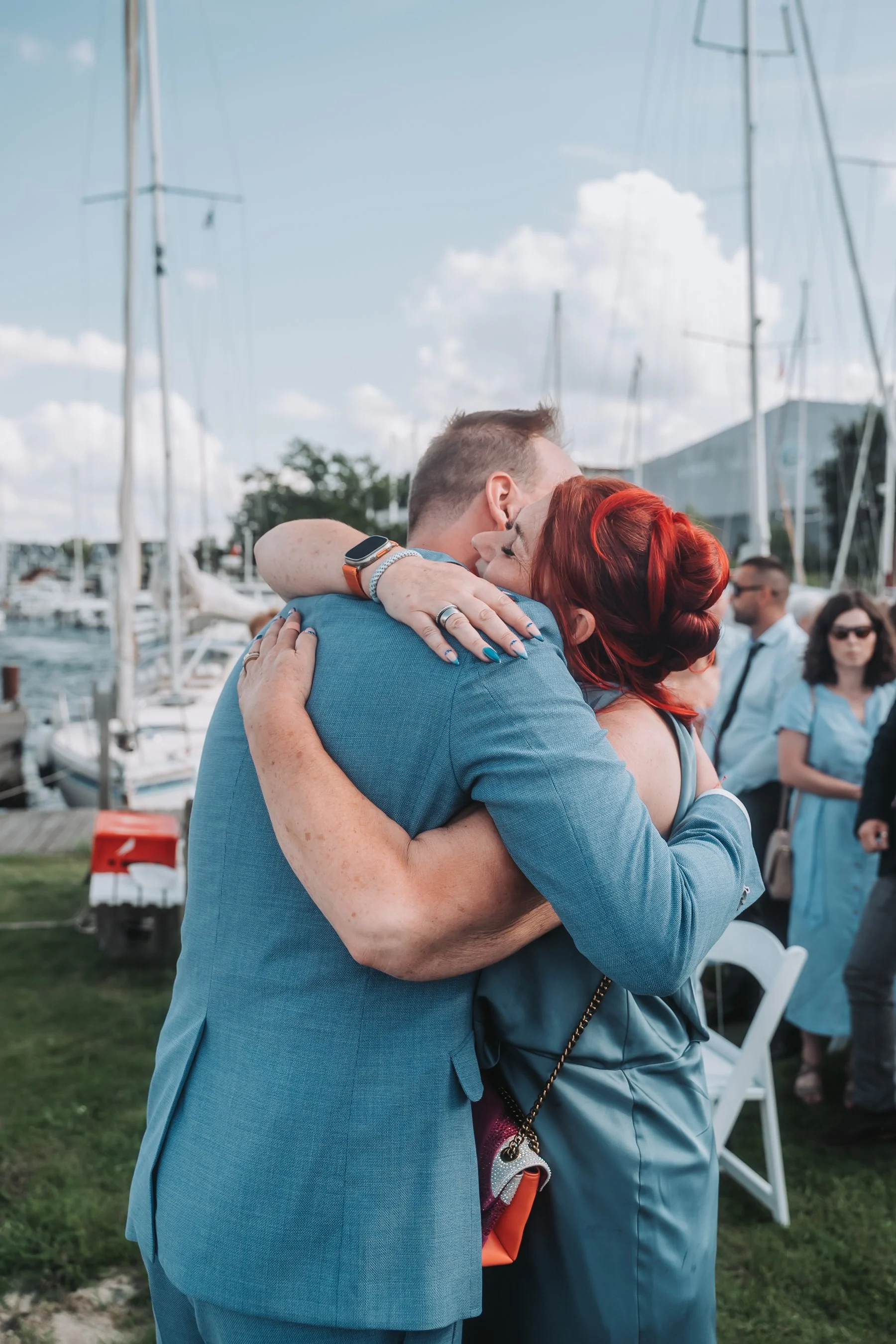 A man and a woman are embracing and hugging each other at an outdoor event near a marina with boats and yachts in the background. The woman has red hair and is wearing a blue dress.