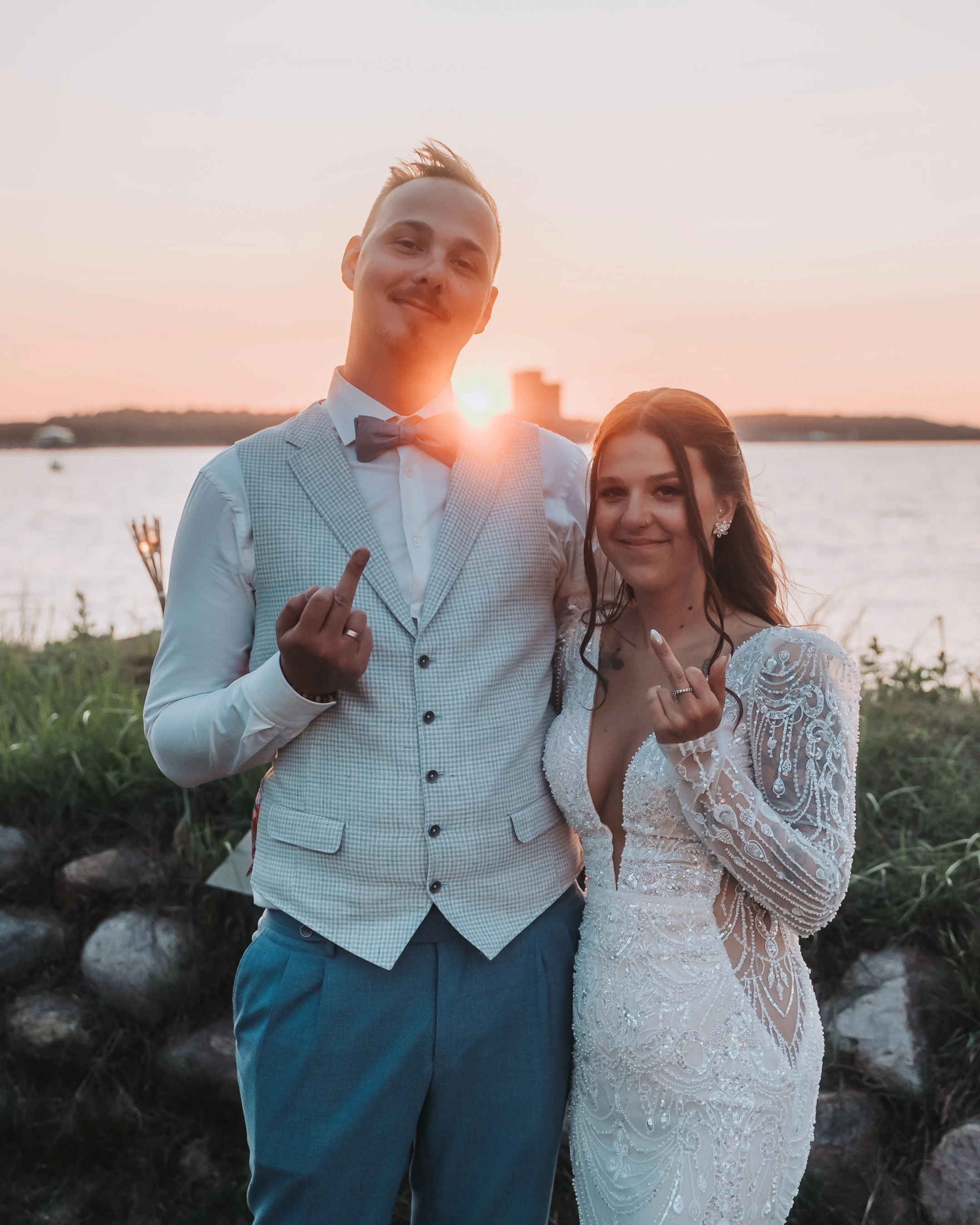 A man in a light-colored suit and a woman in a white wedding dress stand outdoors by a body of water during sunset, both showing their middle fingers and smiling.