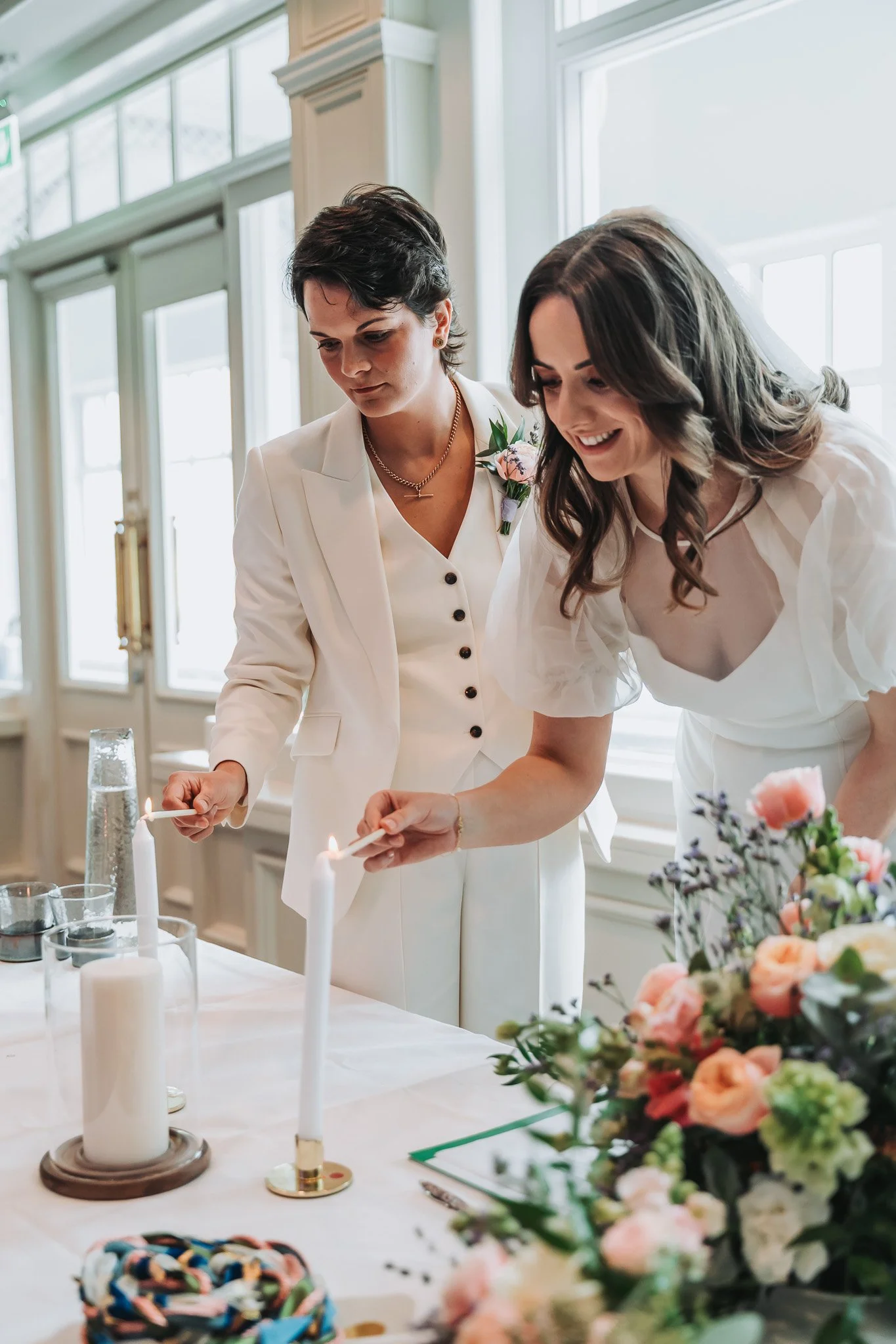 Two women in white dresses and one with a floral boutonniere participate in a candle-lighting ceremony at a table decorated with flowers and candles in a bright room.